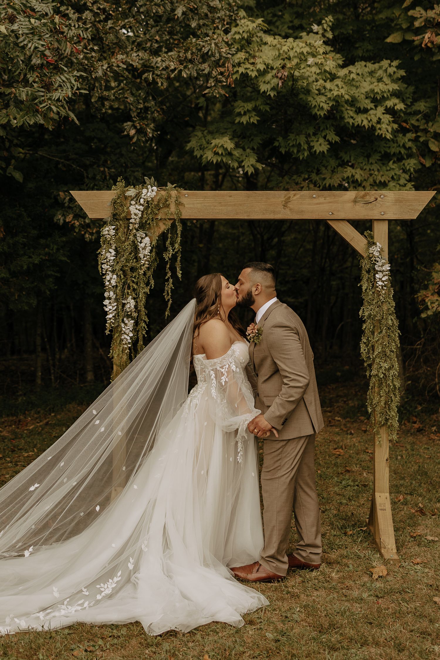 Romantic wedding ceremony moment under a wooden arbor decorated with flowing white fabric and flowers.