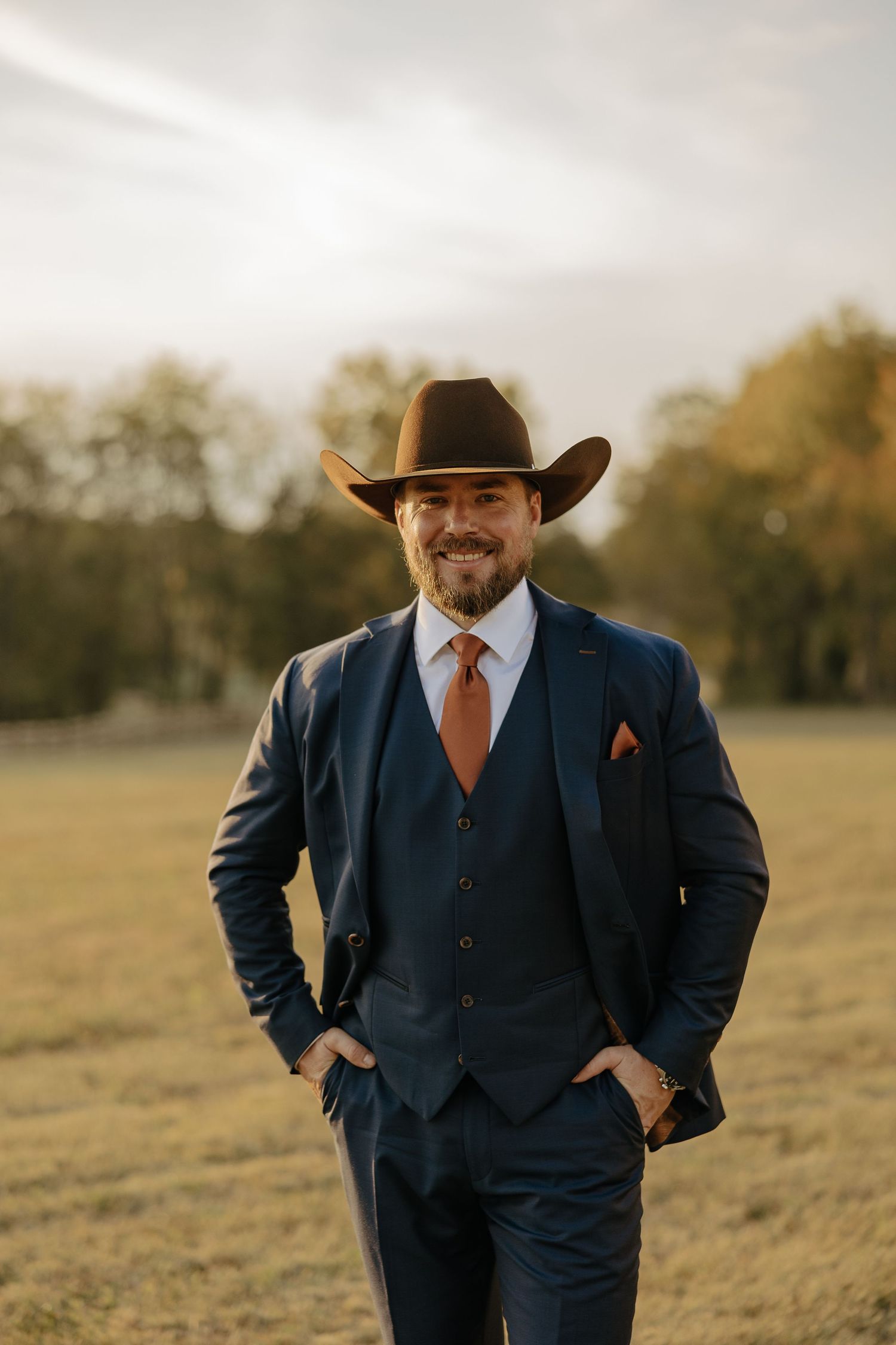 A groom in a navy suit and cowboy hat poses confidently in a golden field at sunset.