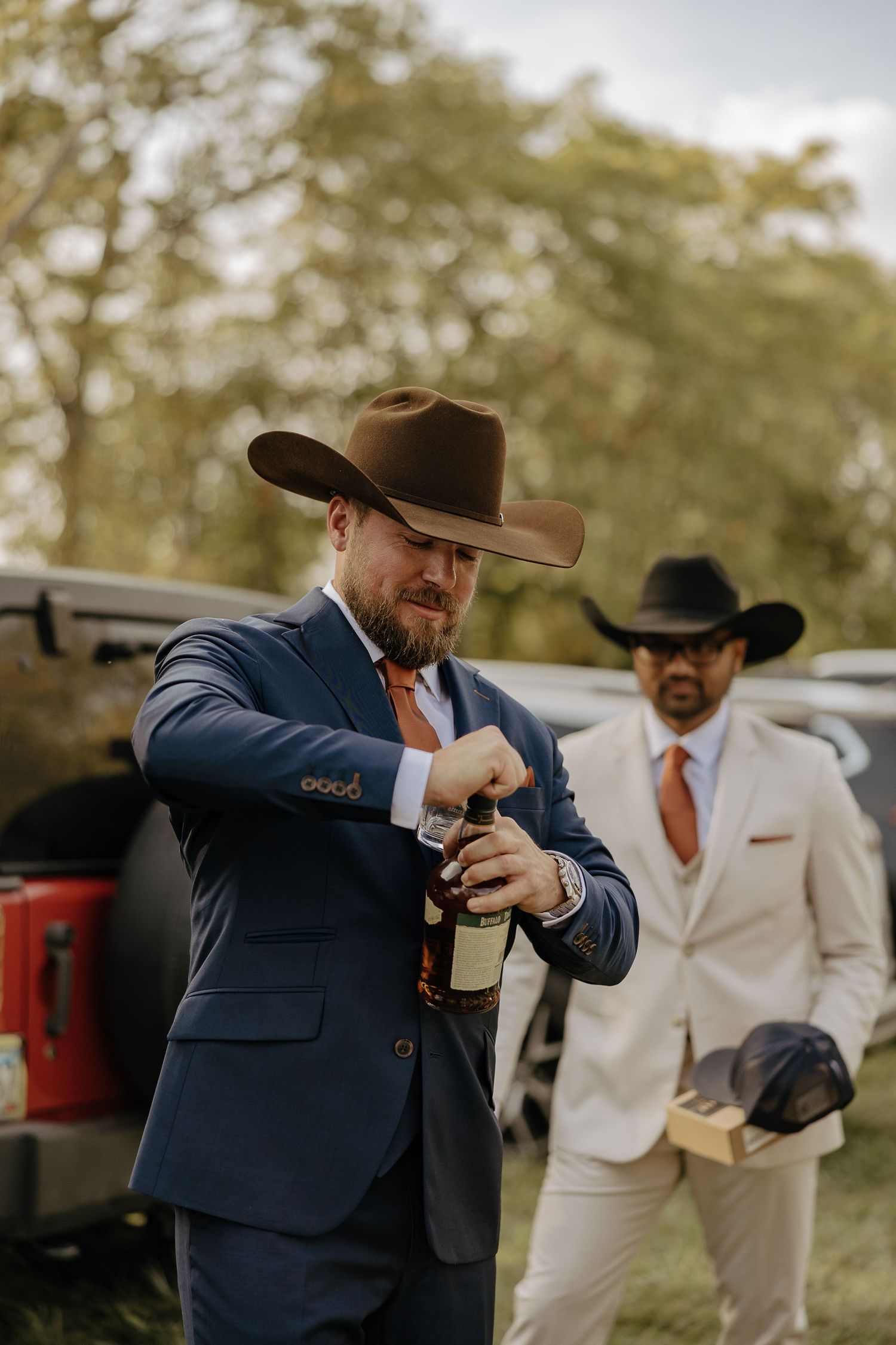 Two people in western attire at an outdoor event, with one in a navy suit opening a bottle.