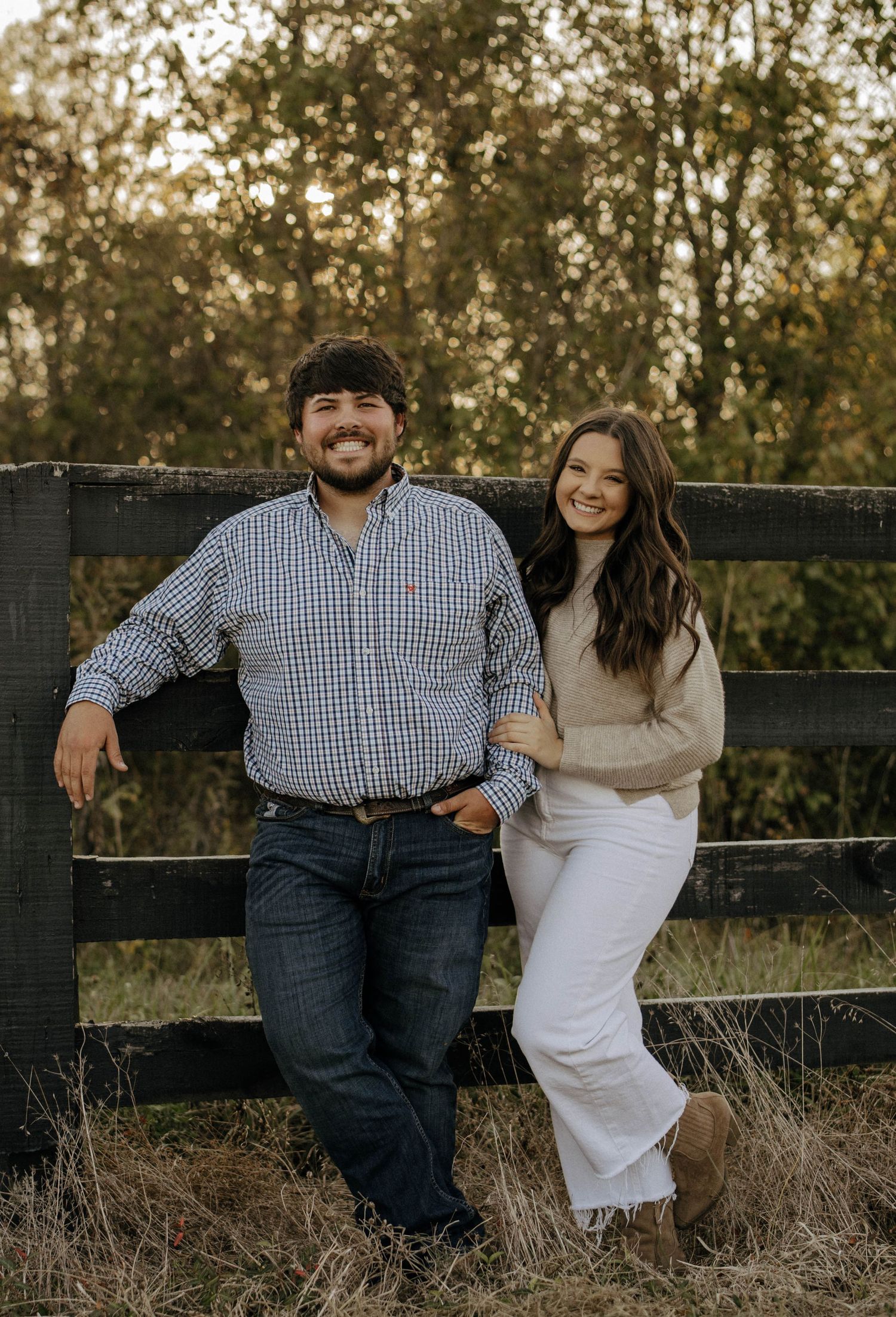 Casual couple relaxing on rustic wooden fence during countryside engagement session.