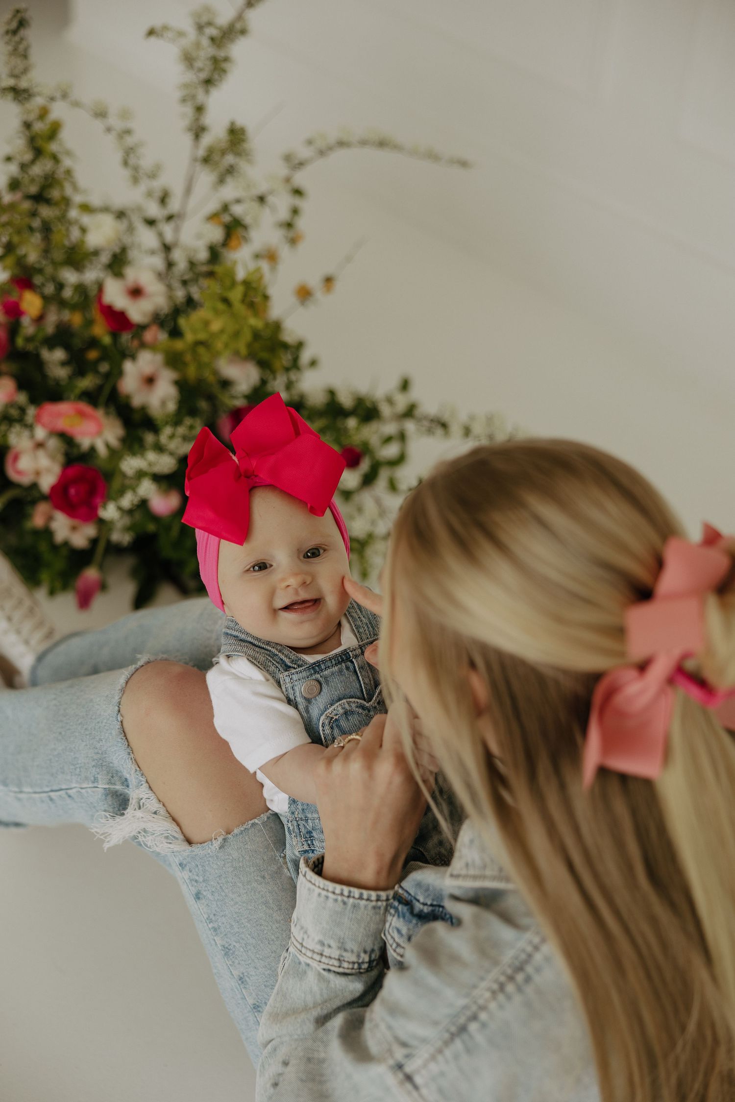 Mother and baby in matching denim outfits play near pink floral display.