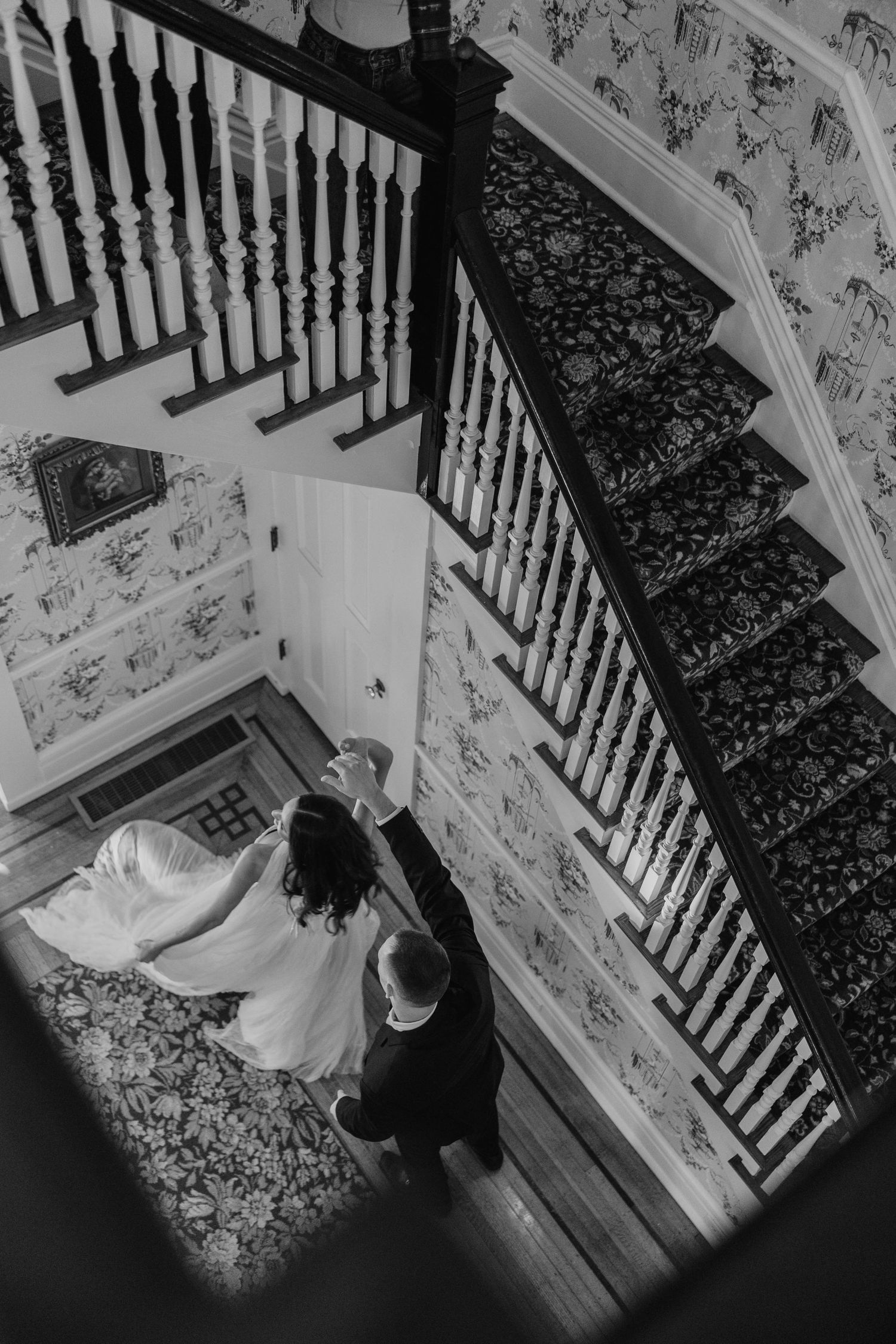Black and white photograph of elegant staircase with ornate carpet runner and floral wallpaper.