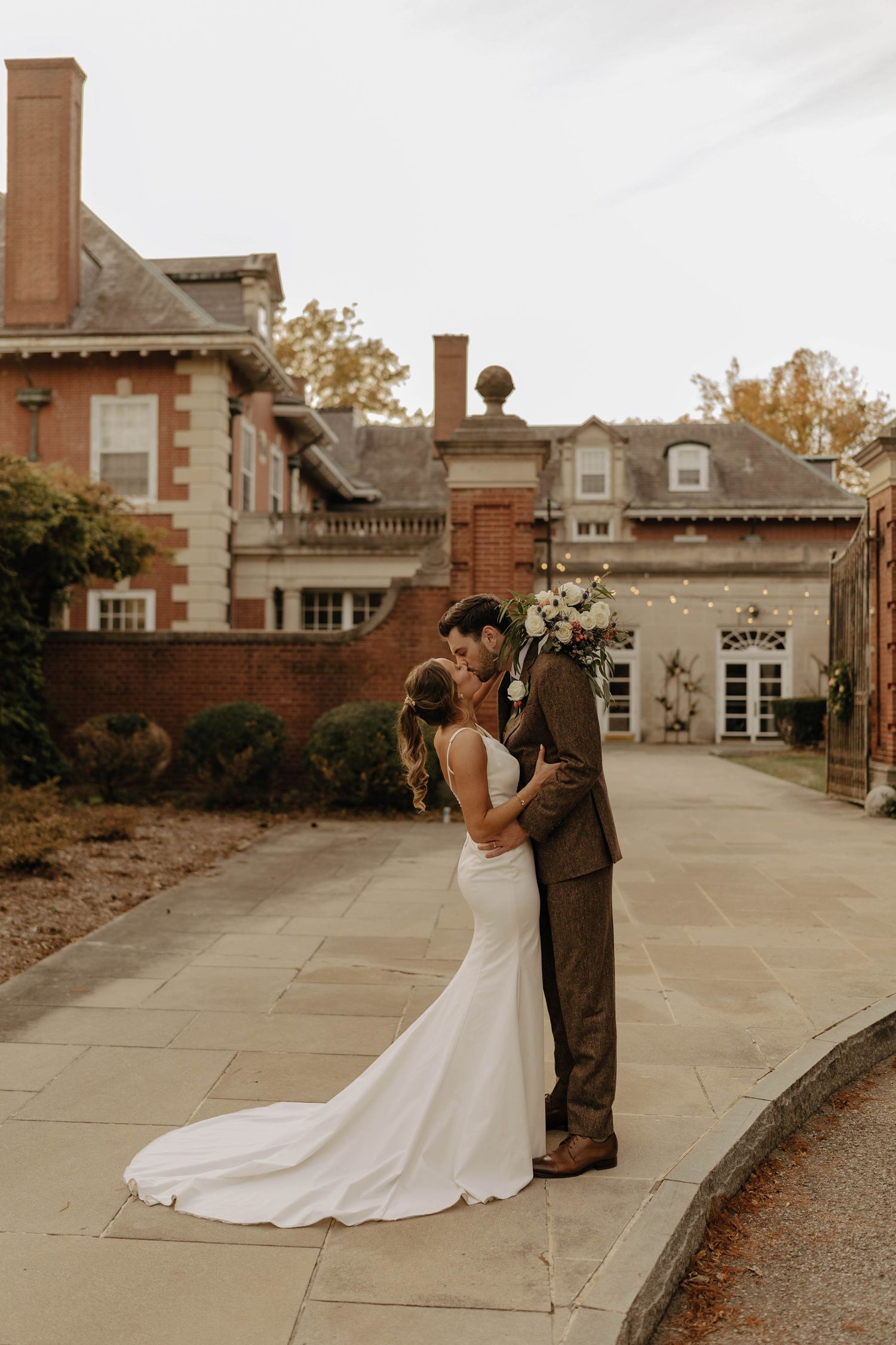 A romantic wedding couple embraces on a stone path outside an elegant brick mansion with autumn foliage in the background.