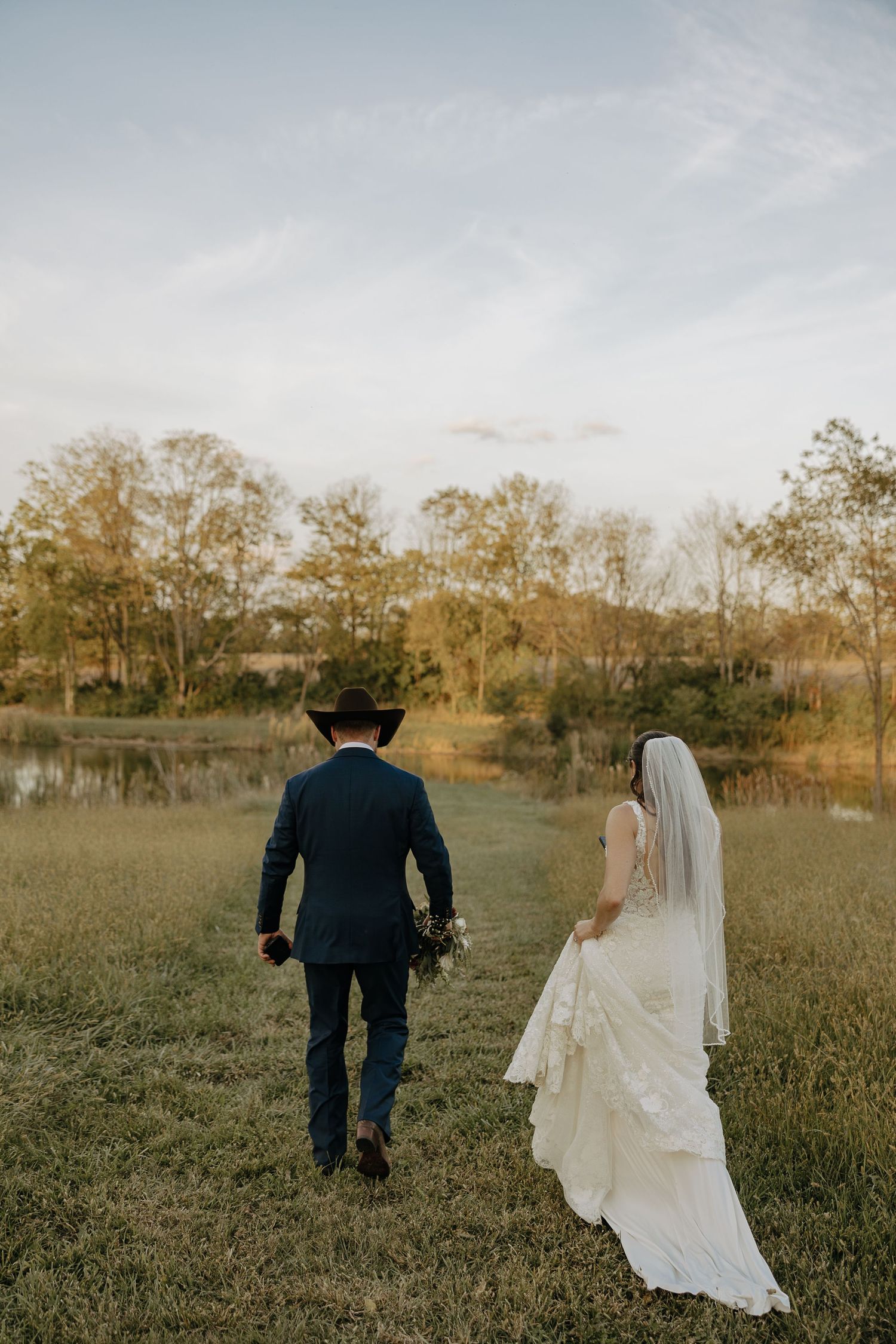 A couple walks through an open field during golden hour with their wedding attire flowing in the breeze.