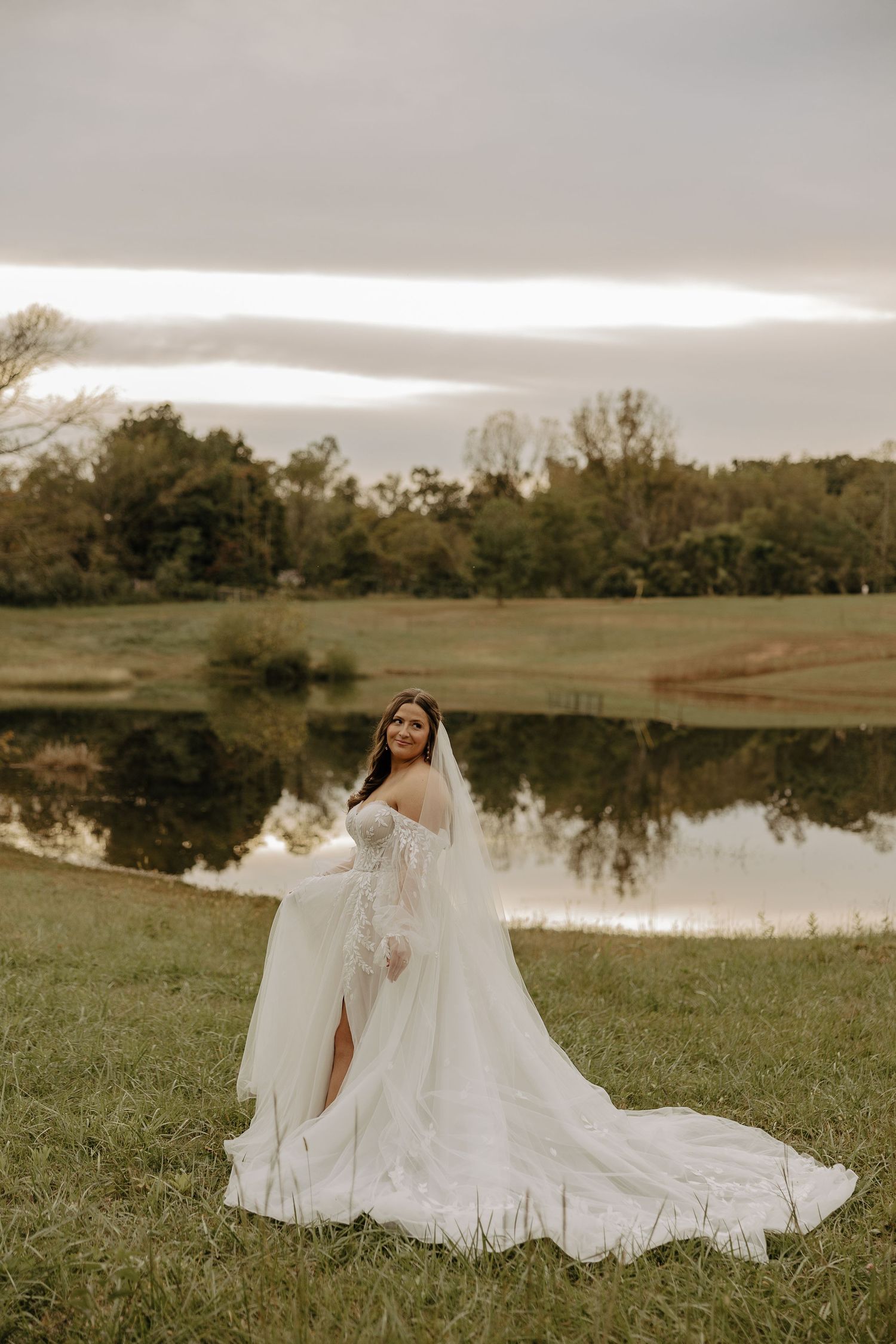 Wedding dress with long train spread out on grass near reflective pond.