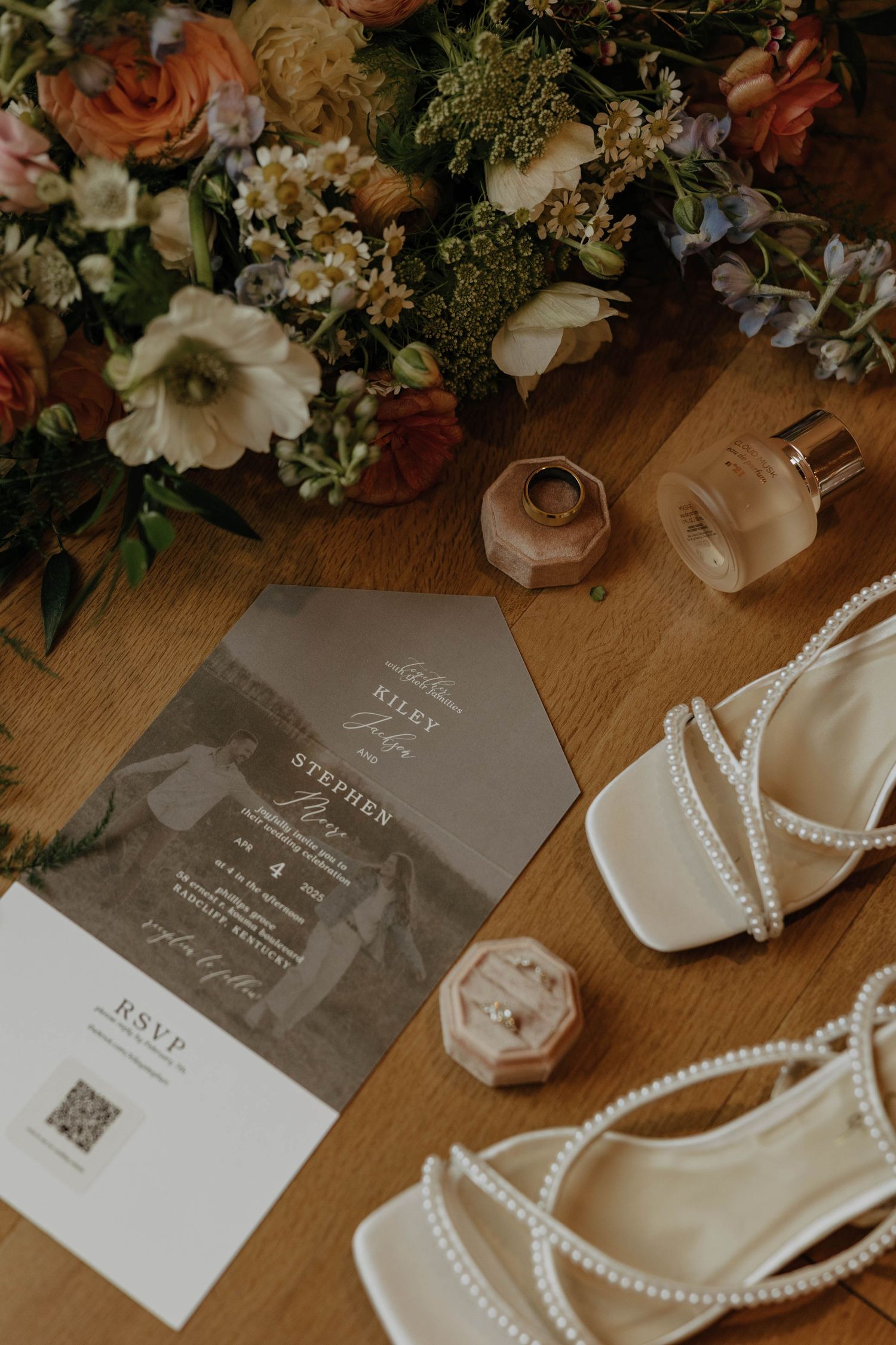 Wedding details including floral arrangements, pearl necklaces, invitation cards and bridal shoes arranged on wooden surface.