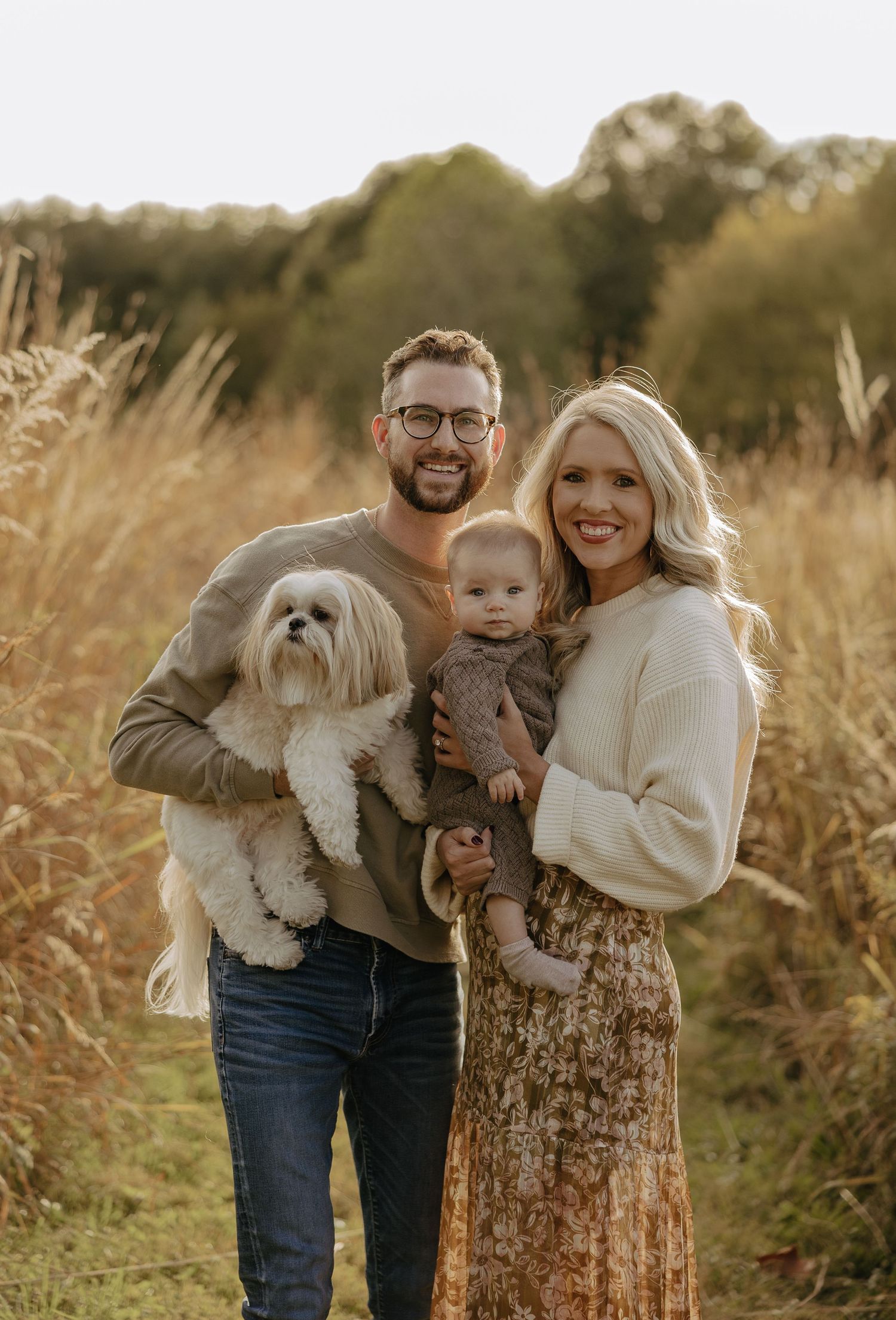 Family portrait in a golden field with a small white dog wearing cozy fall sweaters.