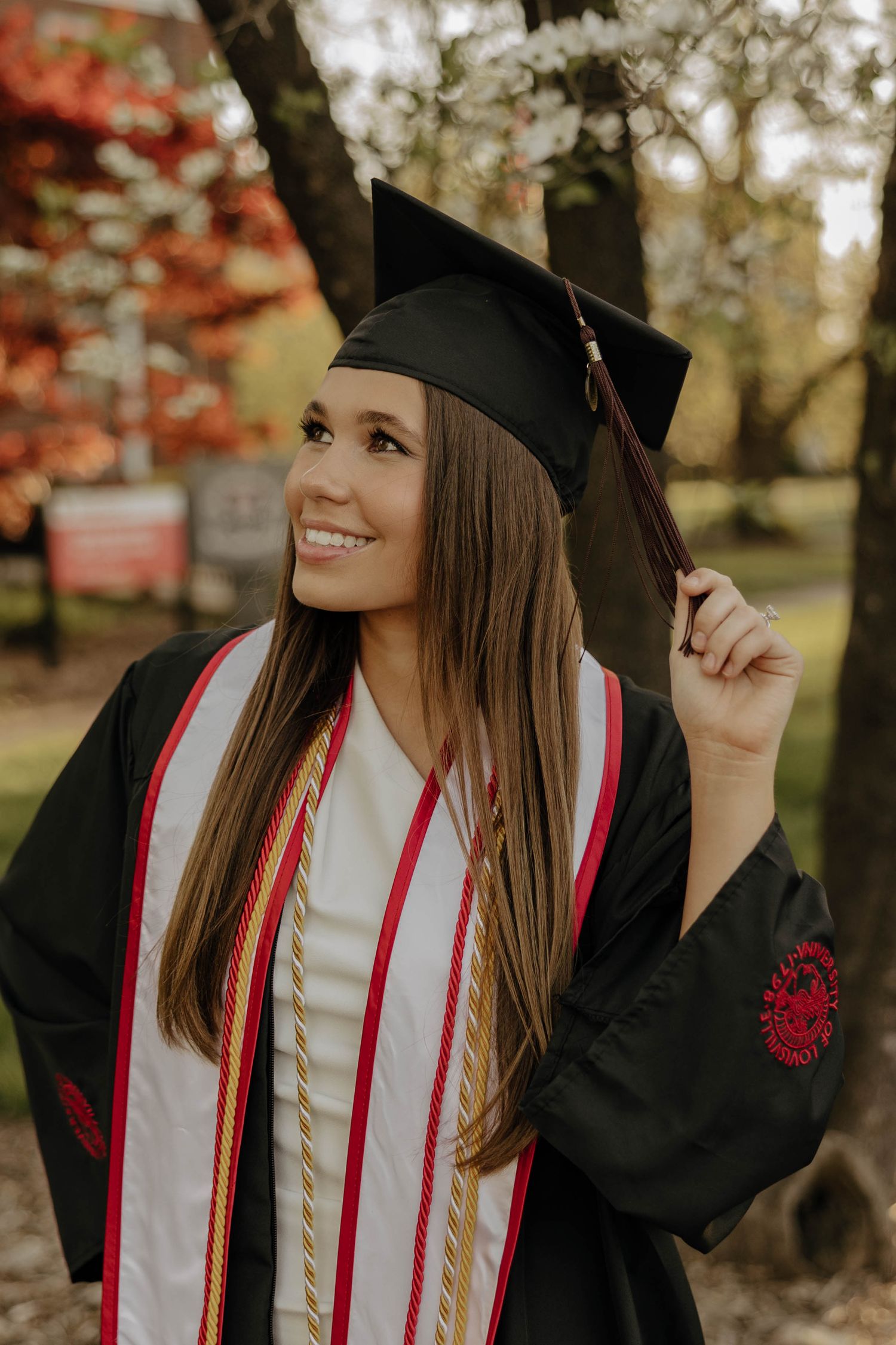 Graduate in academic regalia with red stoles poses against autumn foliage backdrop.