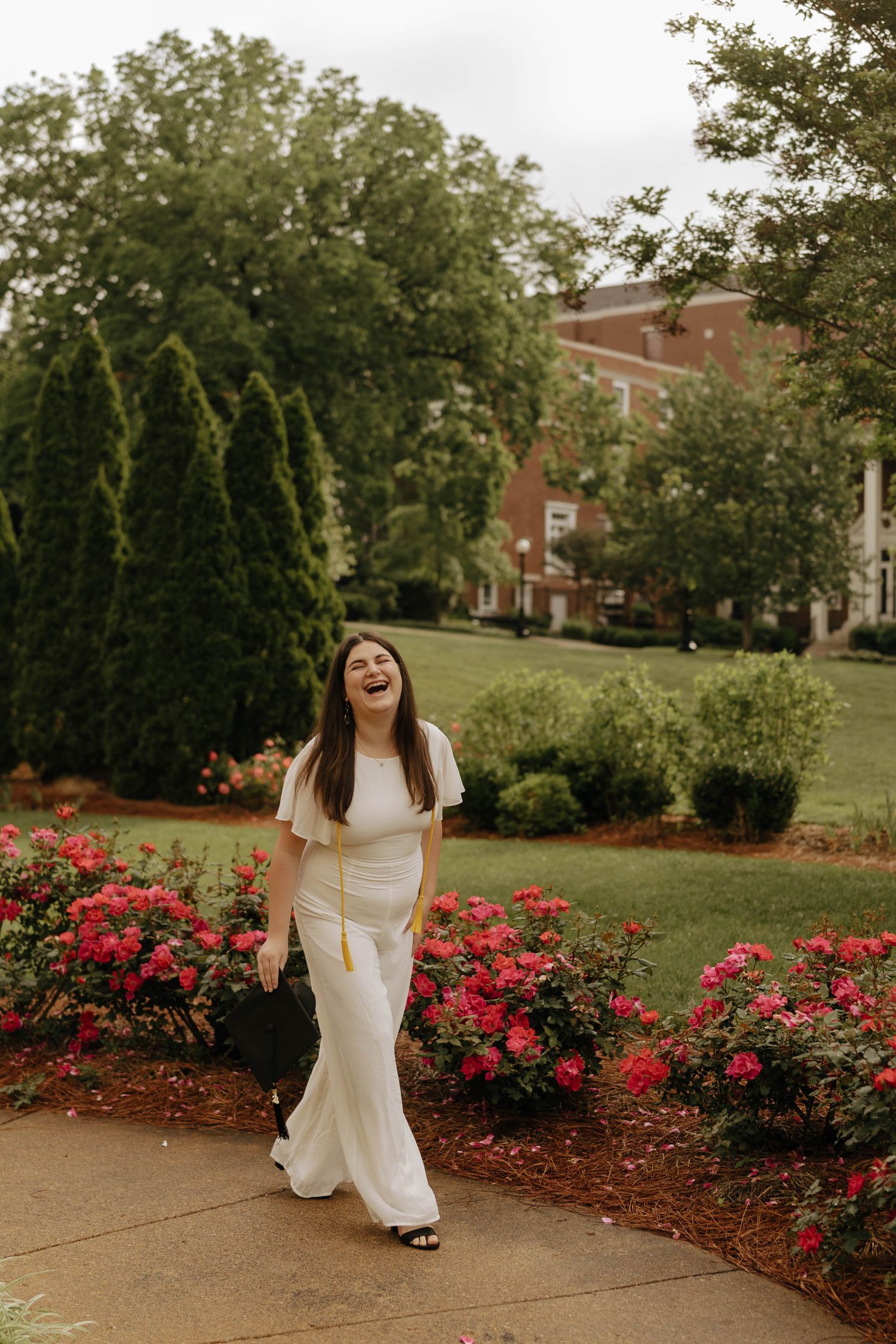 White graduation dress photographed against blooming red rose garden and brick buildings.