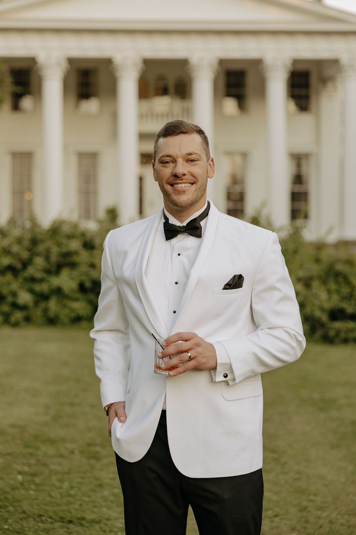 Smiling groom in white tuxedo jacket and black bow tie standing before a grand white columned estate.