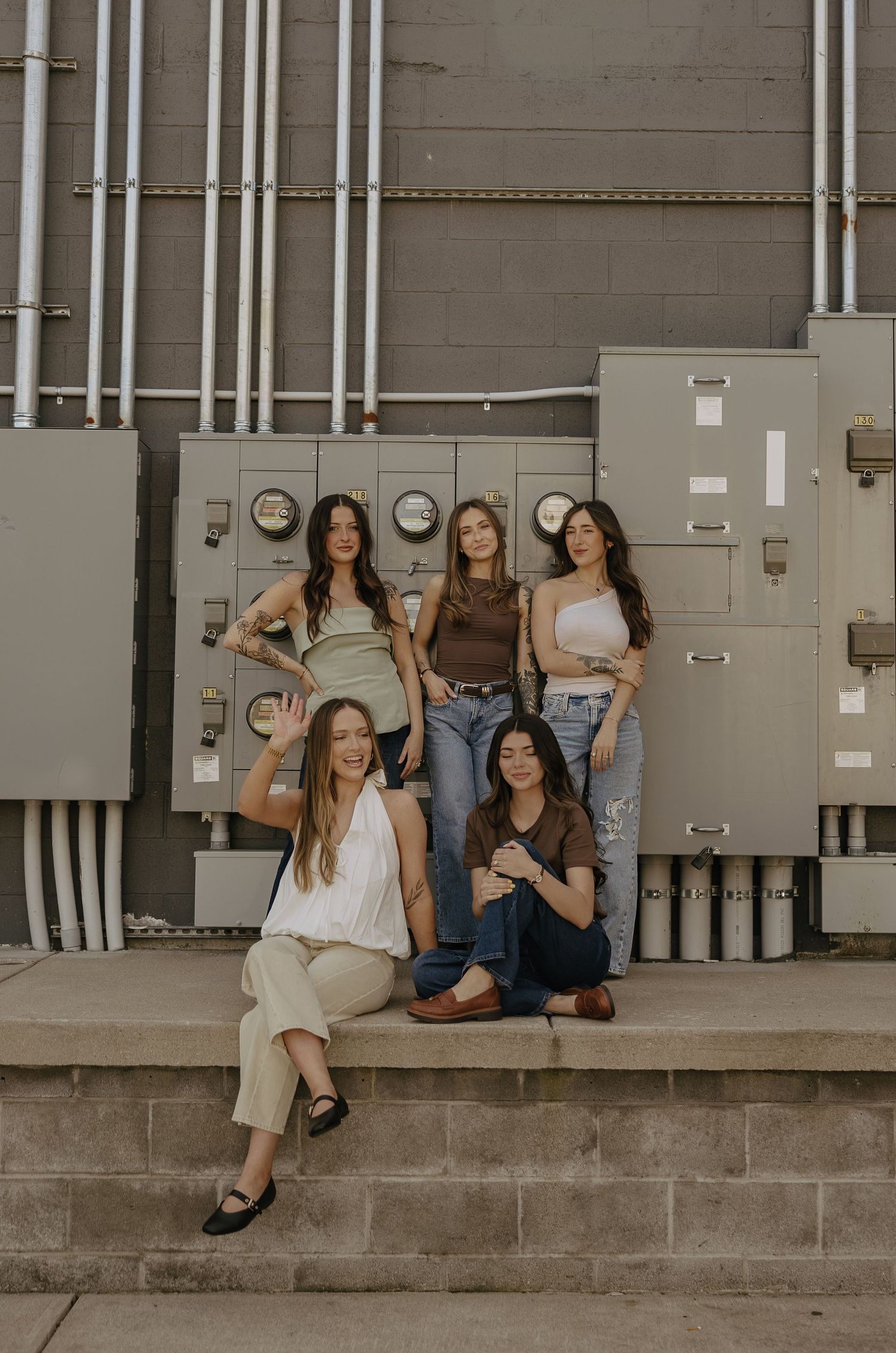 Five young women posing together outdoors near industrial electrical panels, wearing casual trendy outfits.