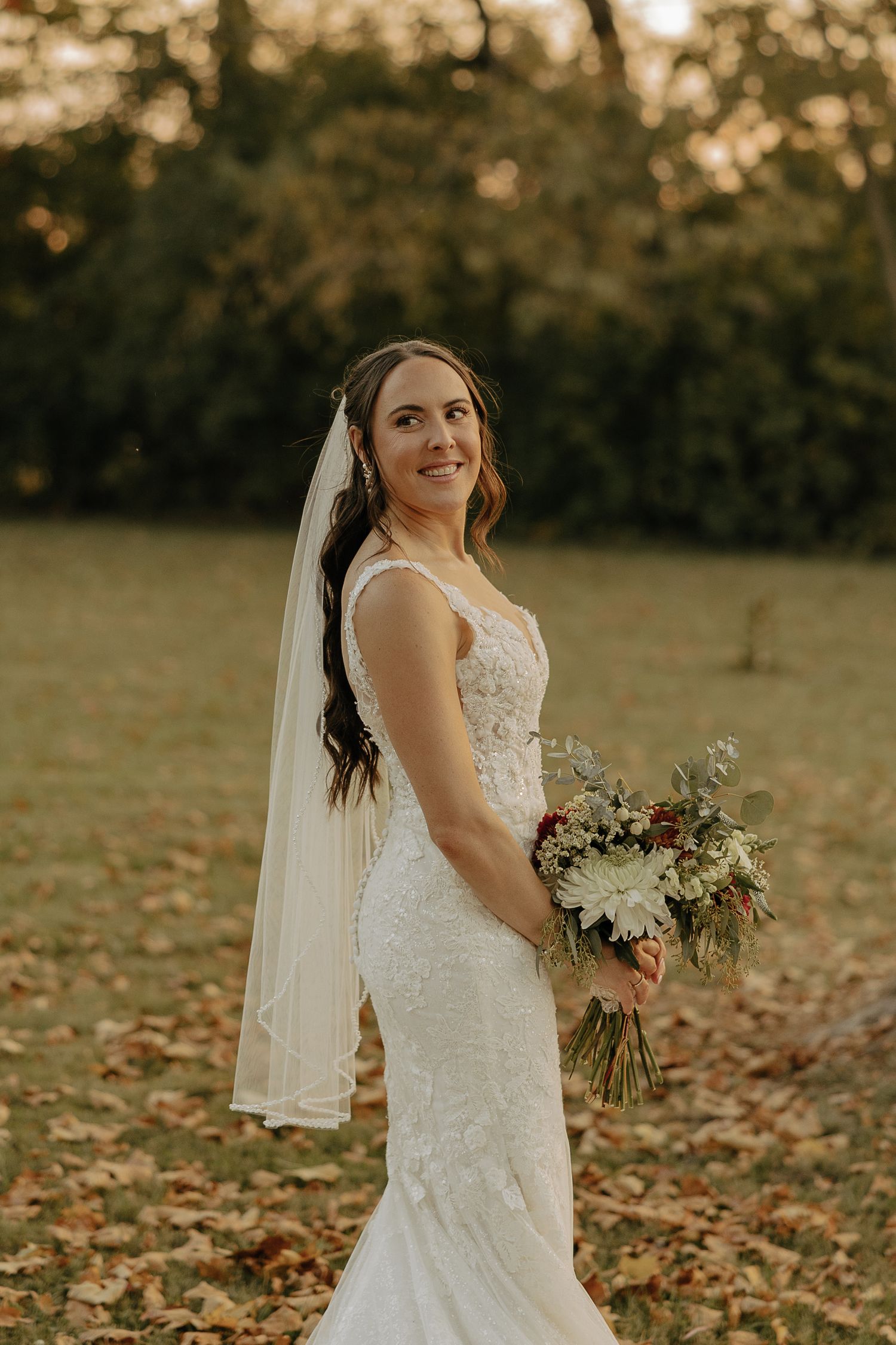 Bride in lace fitted gown holds natural bouquet during autumn outdoor portraits.