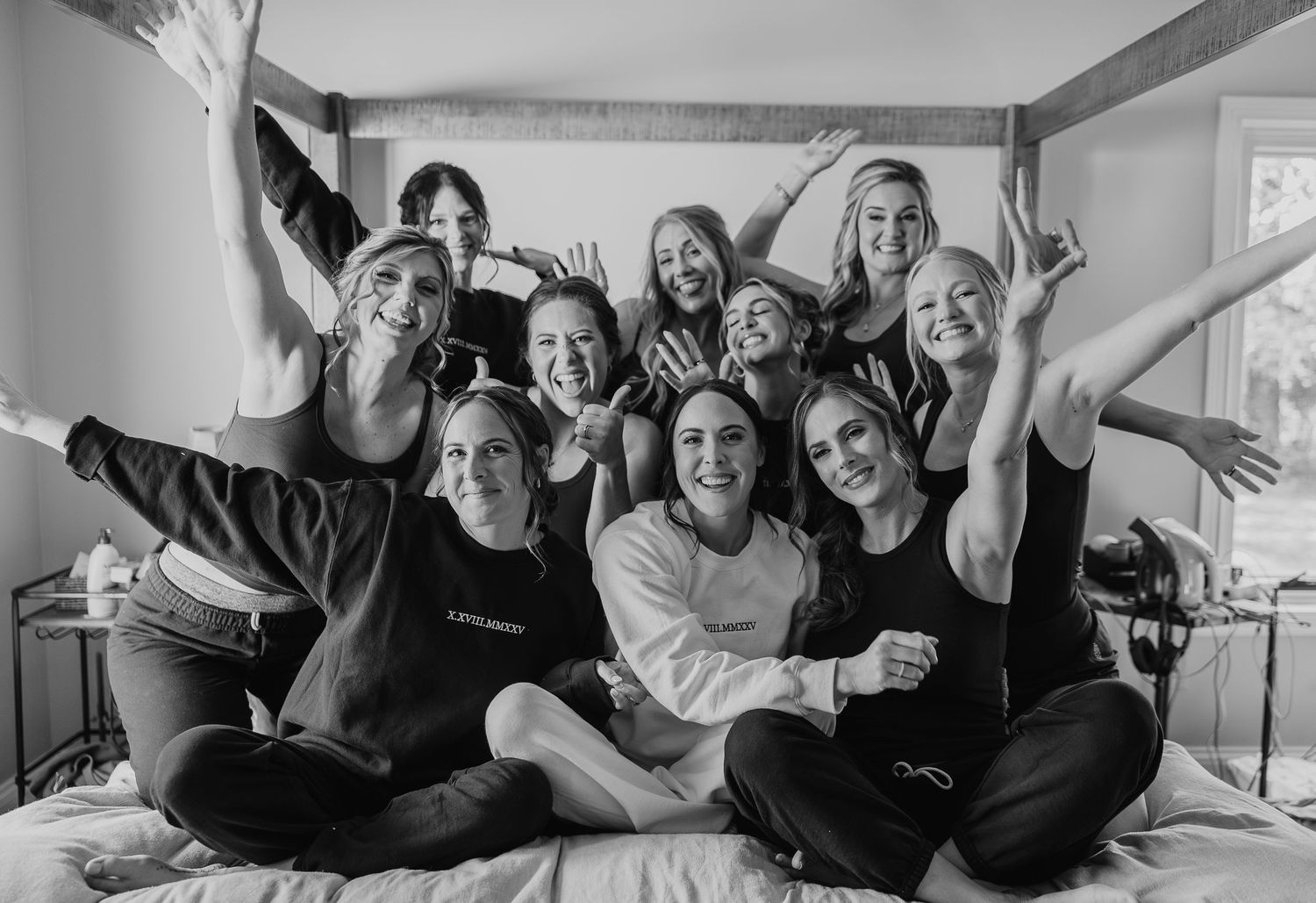 A black and white photo showing a group of people sitting together on a bed, smiling and posing with raised arms.