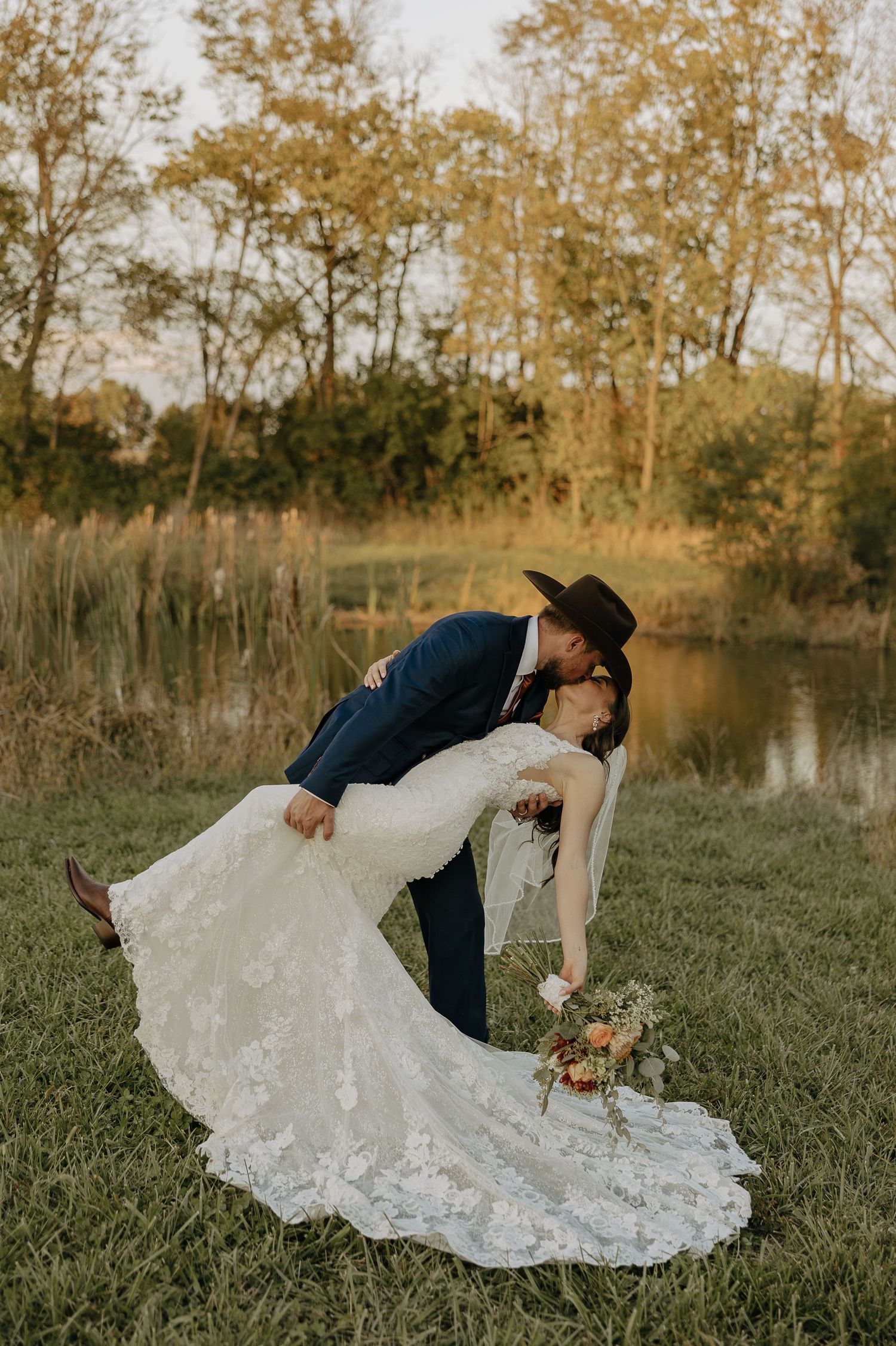 A couple performs a dramatic dip pose in a grassy field with autumn trees in the background.