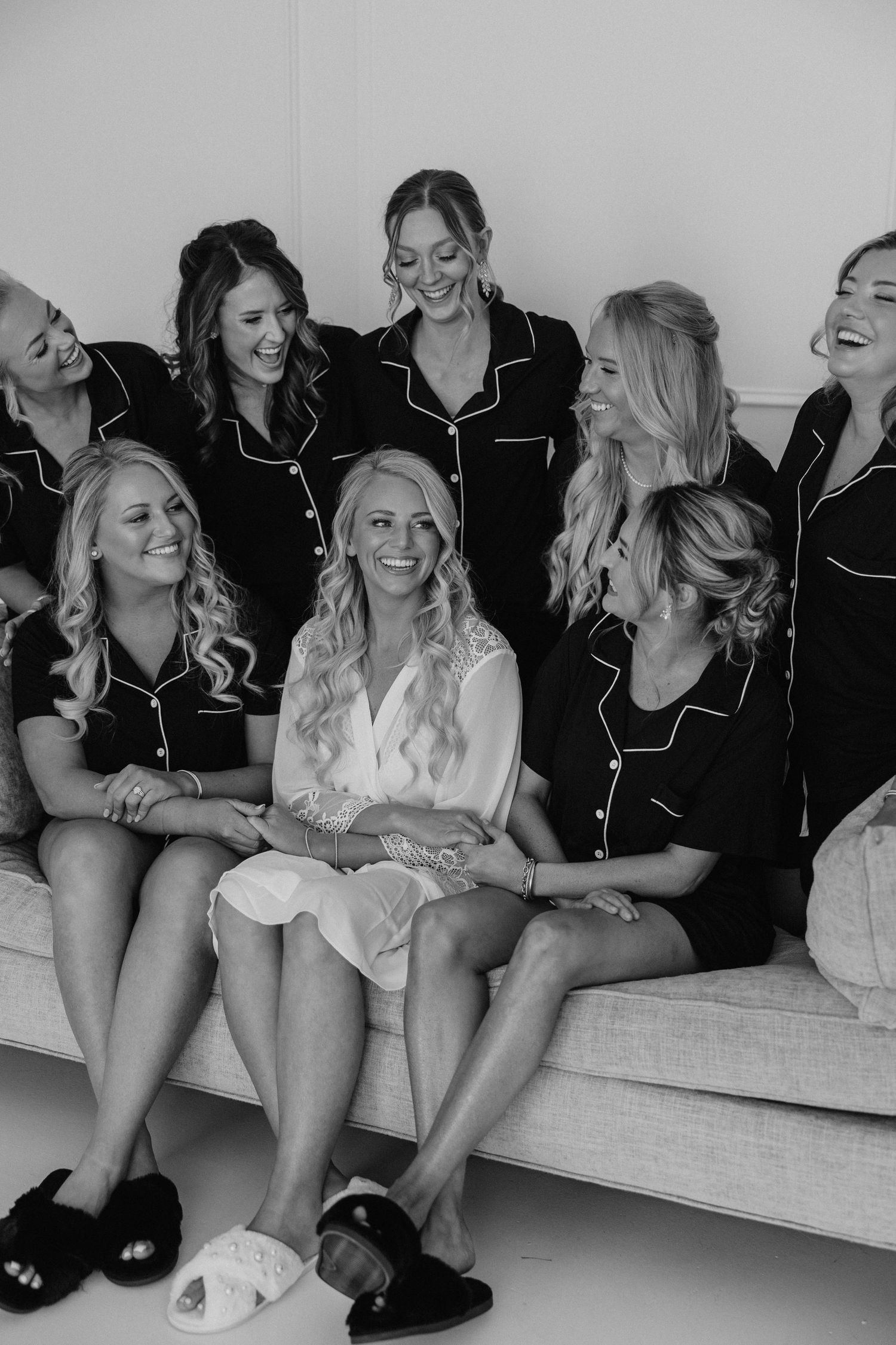 Black and white photo of bridesmaids in matching pajamas laughing together on a couch during wedding preparations.