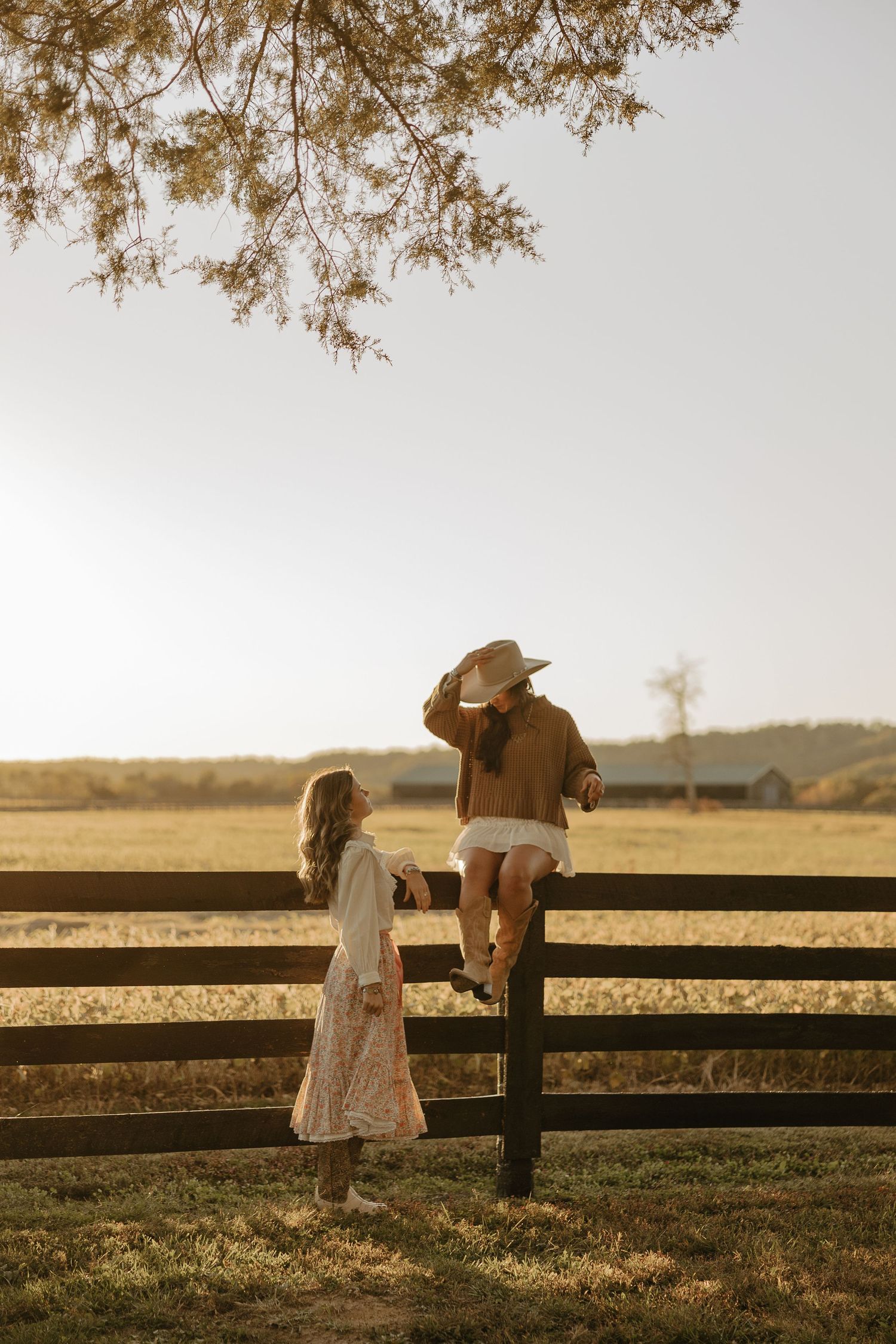 Romantic countryside scene with two people sitting on a wooden fence at sunset.