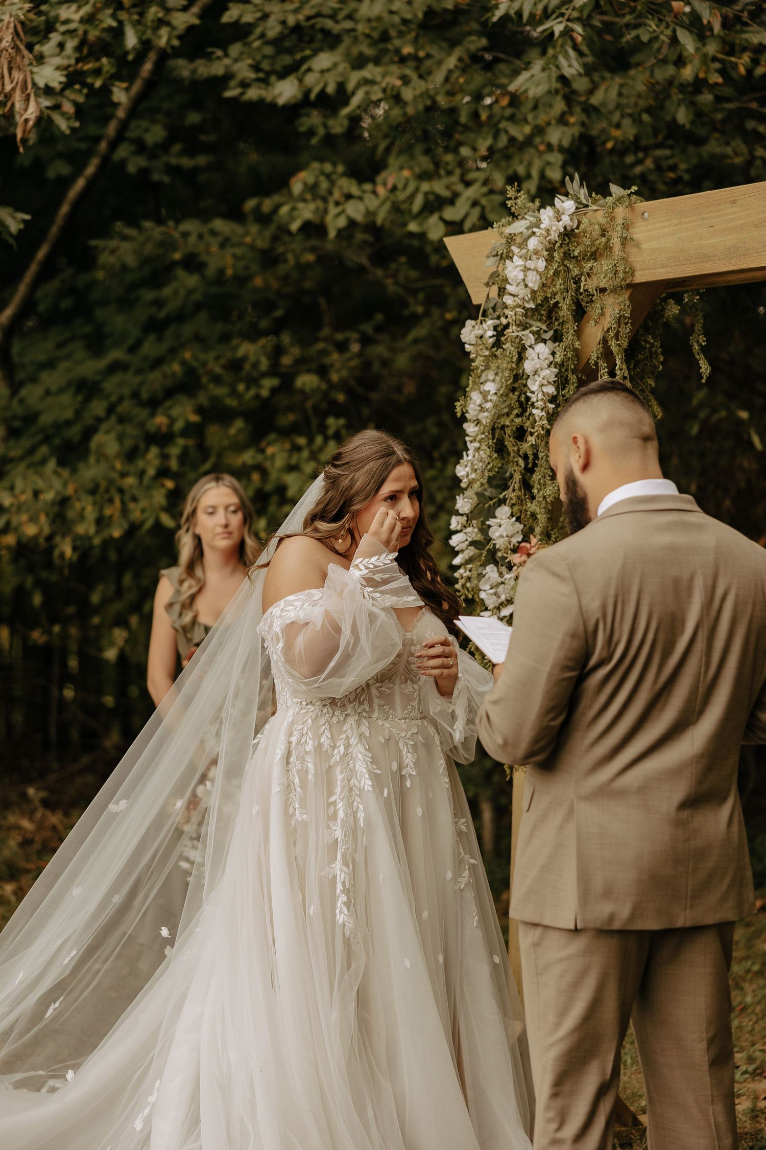 A couple exchanges vows under a wooden arch decorated with white flowers during an outdoor forest wedding ceremony.