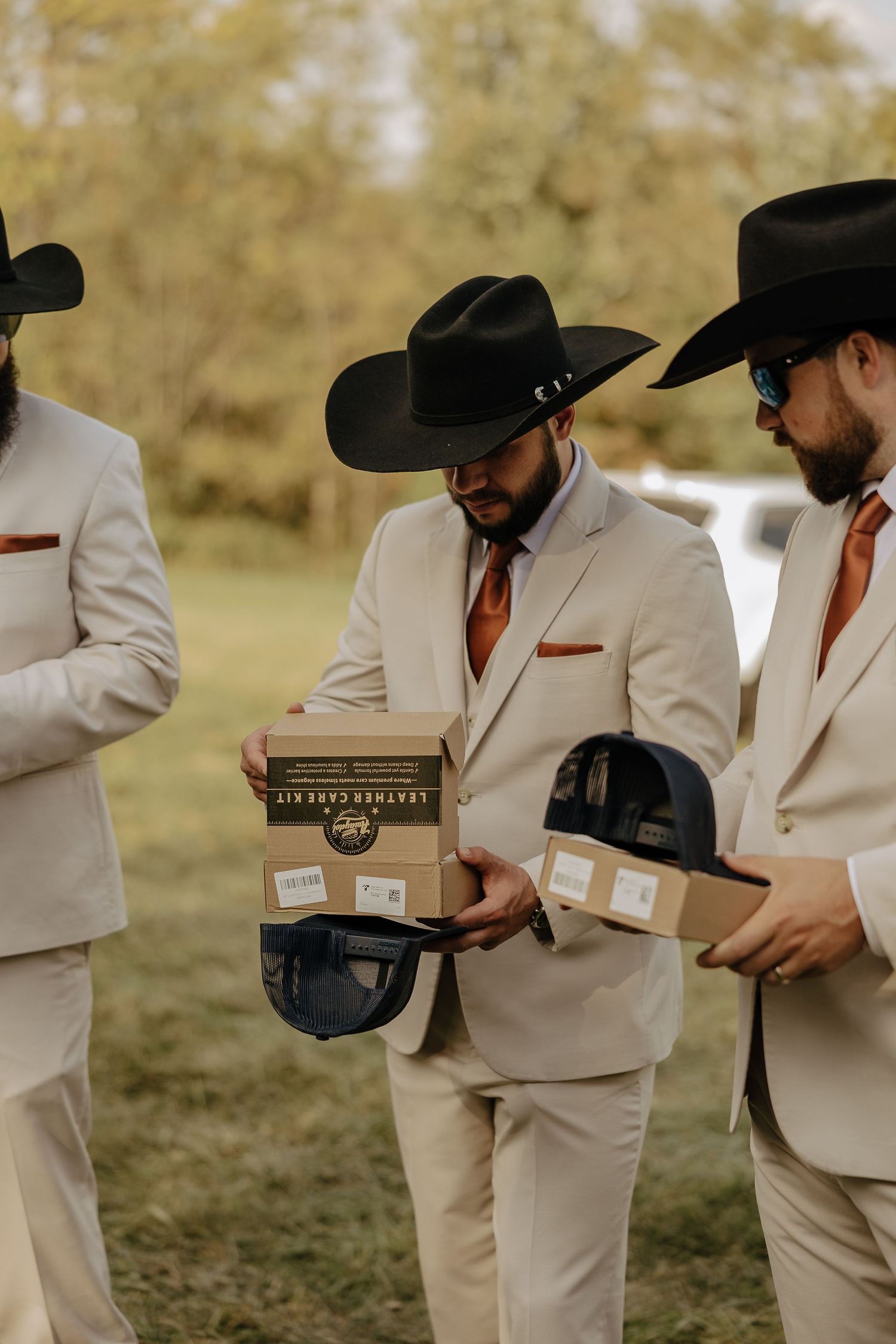Groomsmen in light colored suits and black cowboy hats stand in a row outdoors.