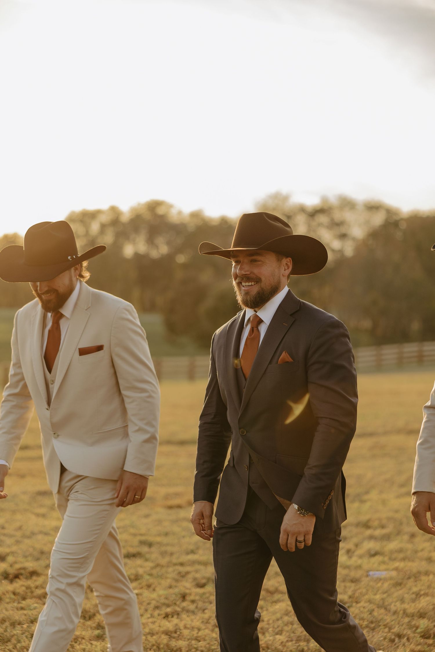 Groomsmen in cowboy hats and suits walk across a sunlit field during golden hour.