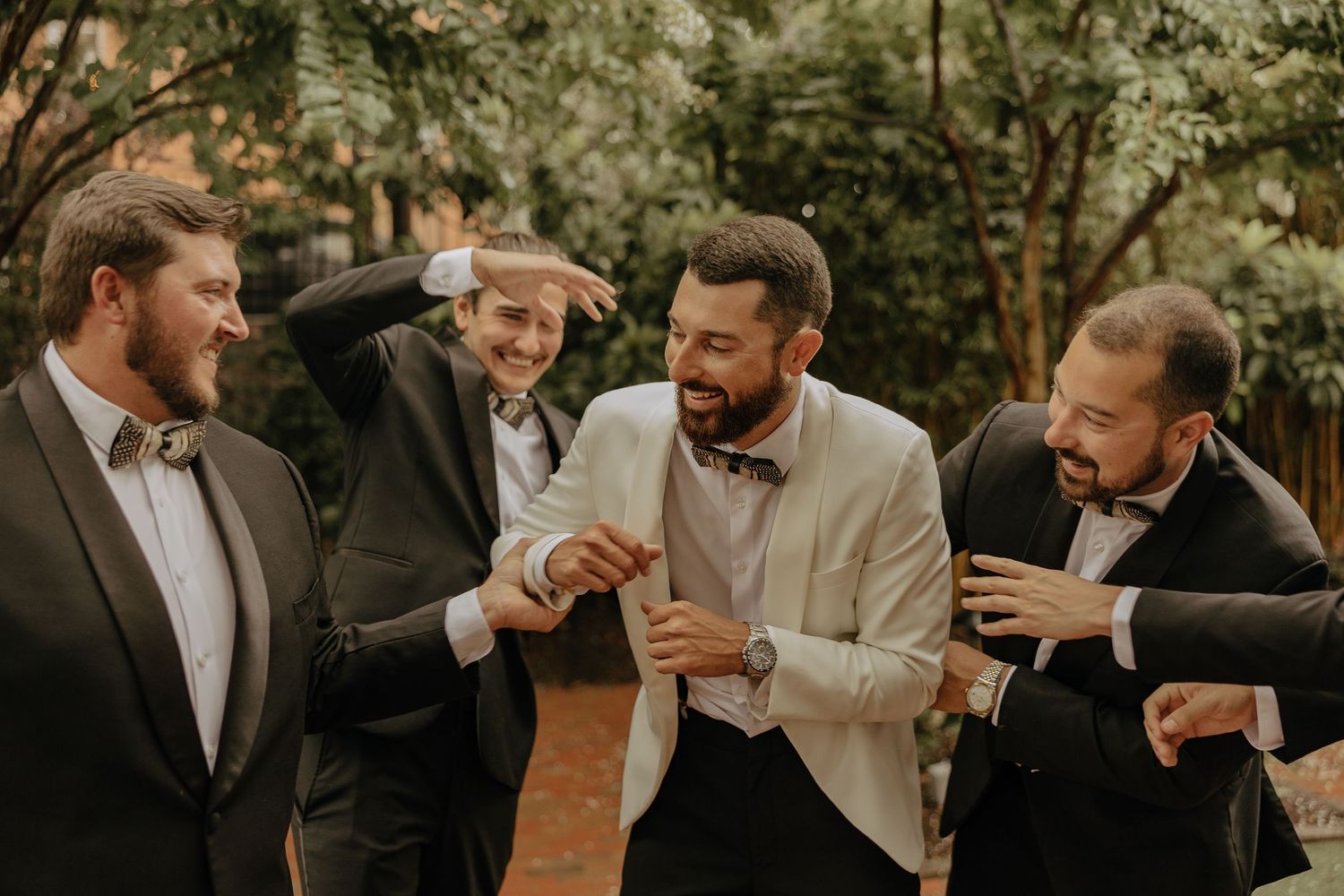 A group of groomsmen share a lighthearted moment laughing together outdoors in formal attire.