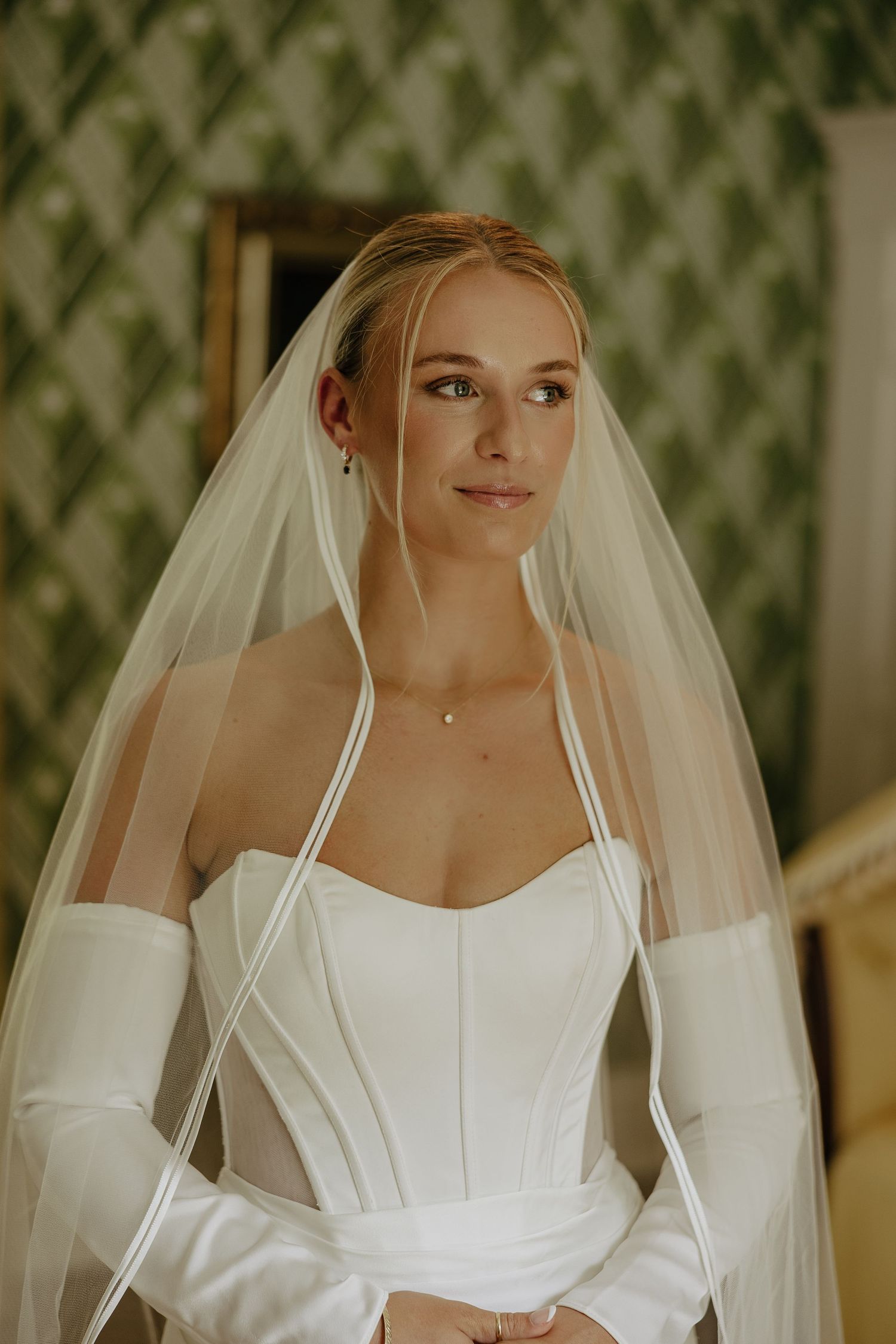 Bride in white strapless gown and satin-edged veil poses elegantly before green wallpaper.