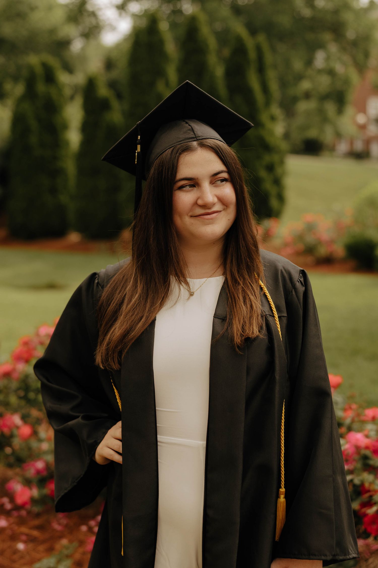 Graduate in black cap and gown with gold tassel poses near flowering garden.