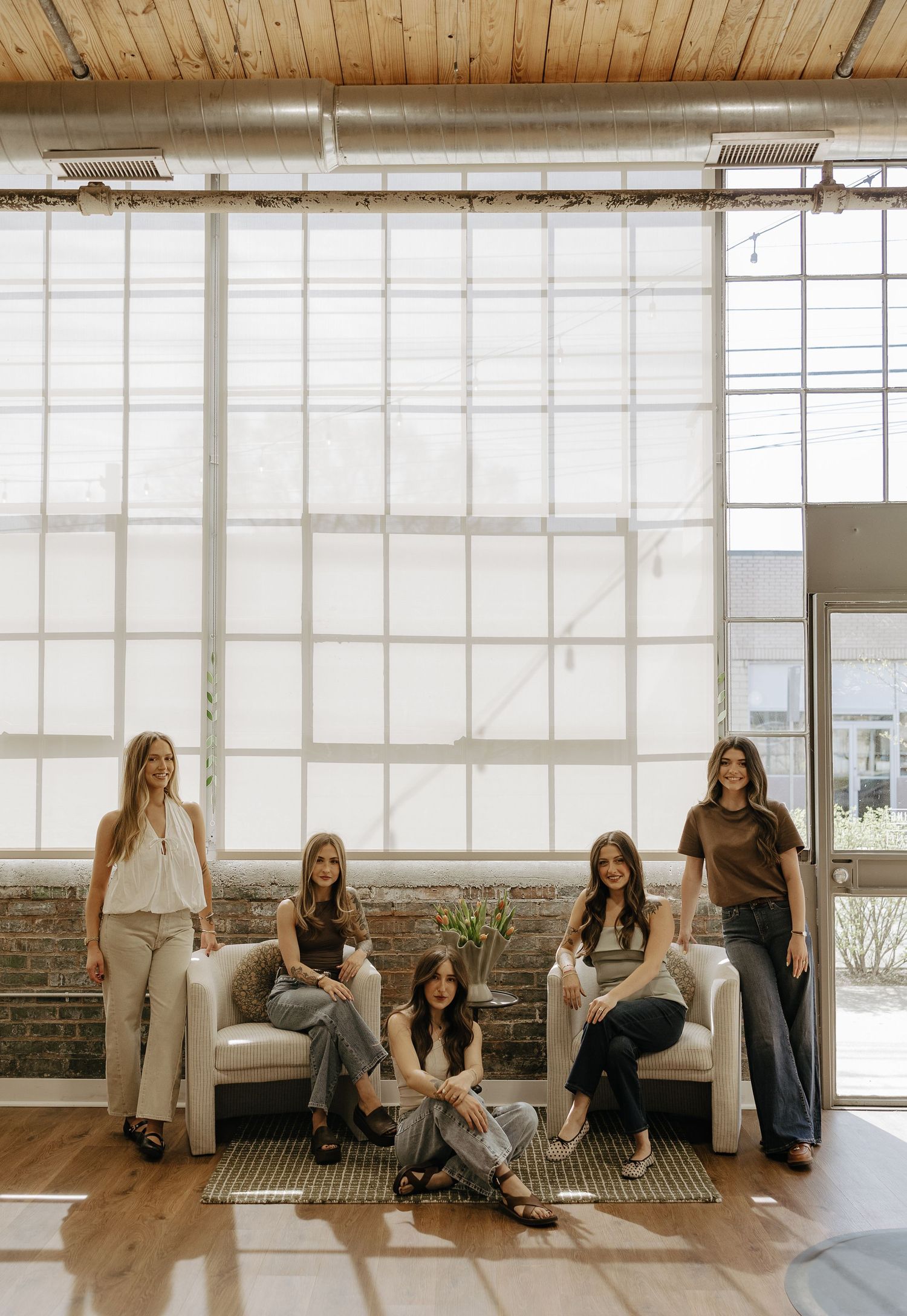 Five stylish women pose in a bright industrial loft with large windows, exposed brick, and modern white chairs.