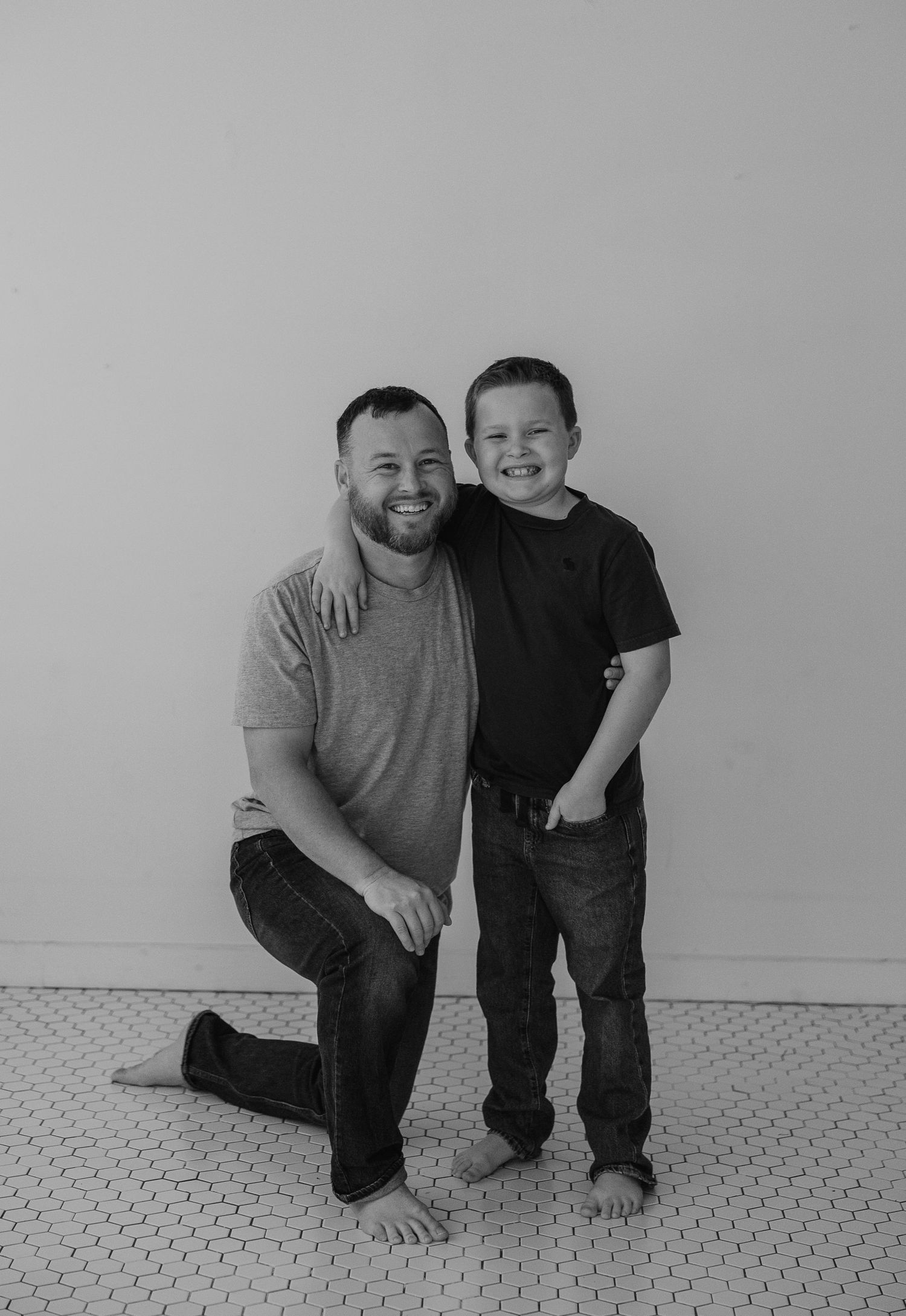 A parent and child pose together against a gray wall in a casual black and white portrait.