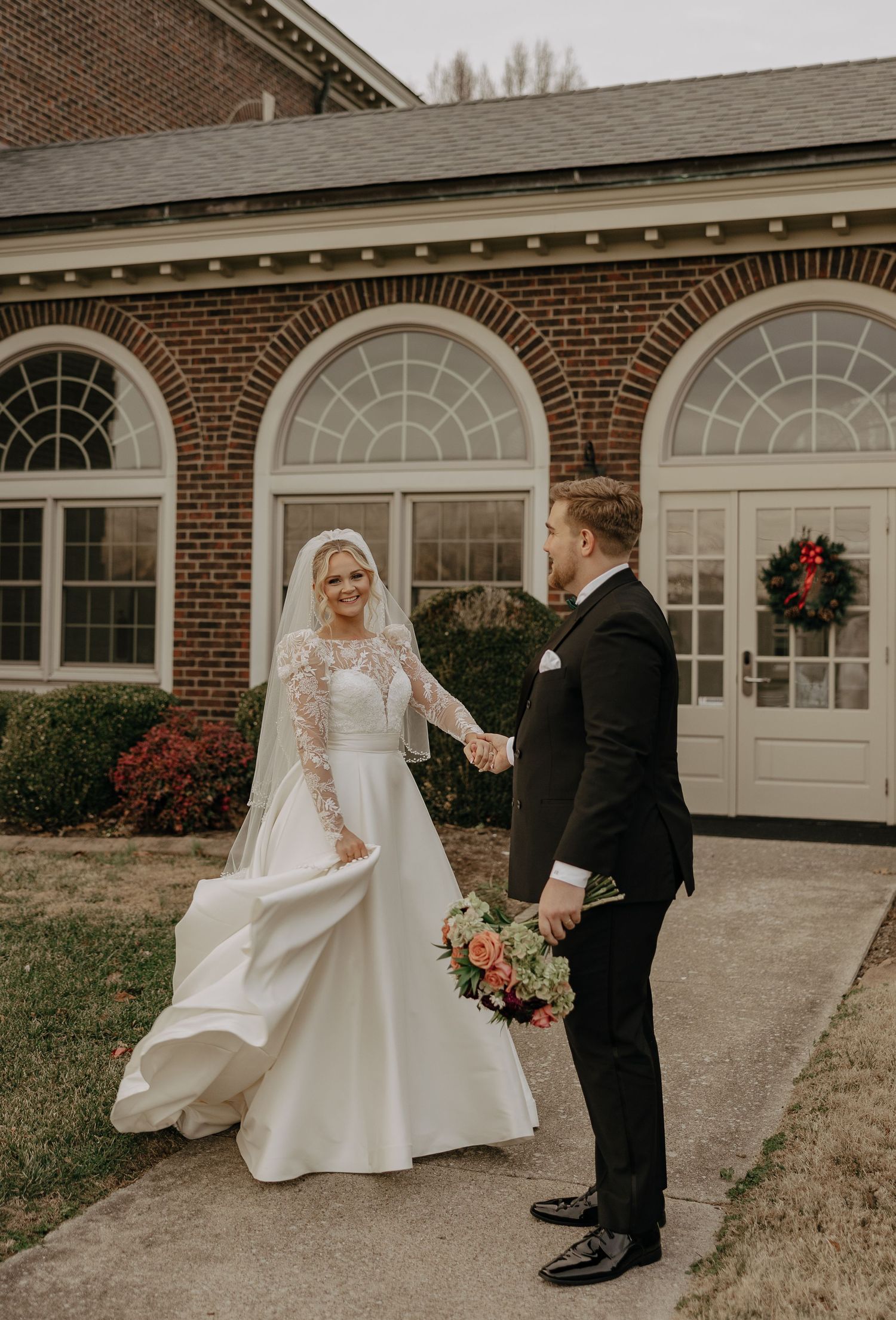 A bride in a flowing white wedding dress twirls and dances with her groom outside a brick building.