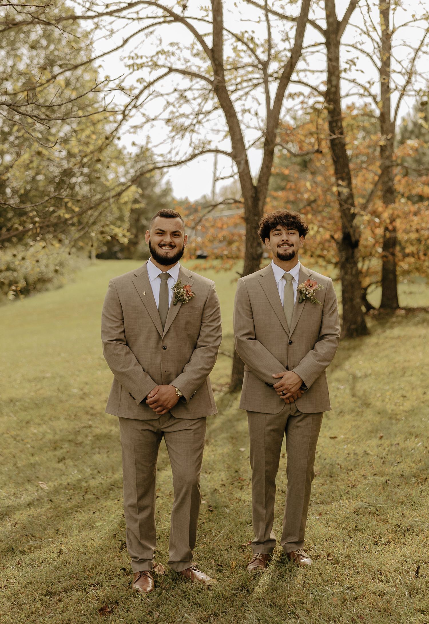 Groomsmen in tan suits standing together on a grassy hillside with autumn trees in the background.