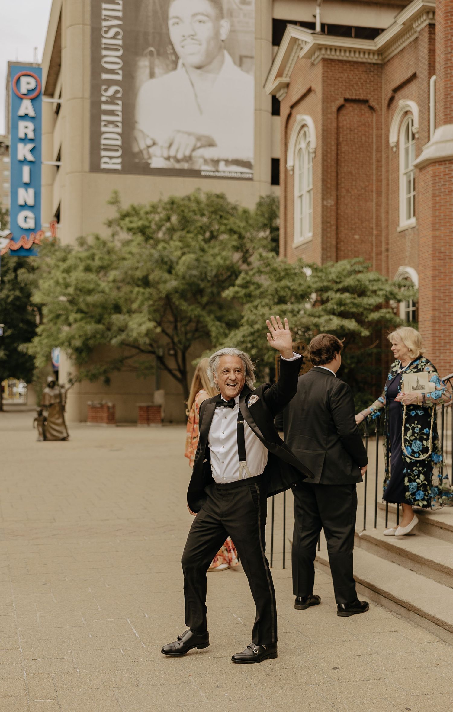 Gentleman in tuxedo waves cheerfully outside Rudells Louisville venue during wedding celebration.
