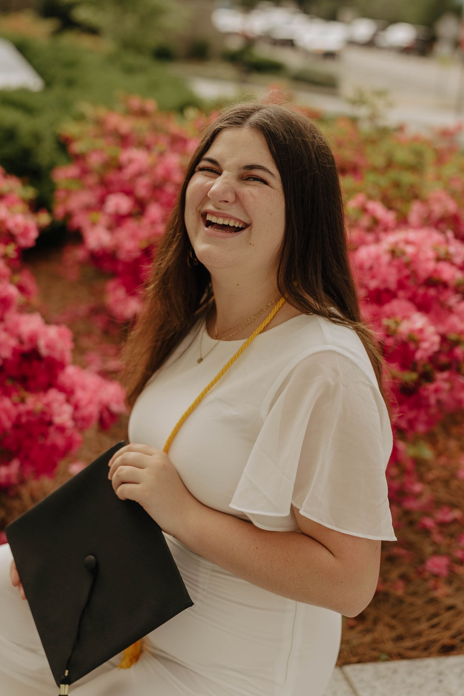 Graduate celebrates with cap against blooming pink azalea bushes.
