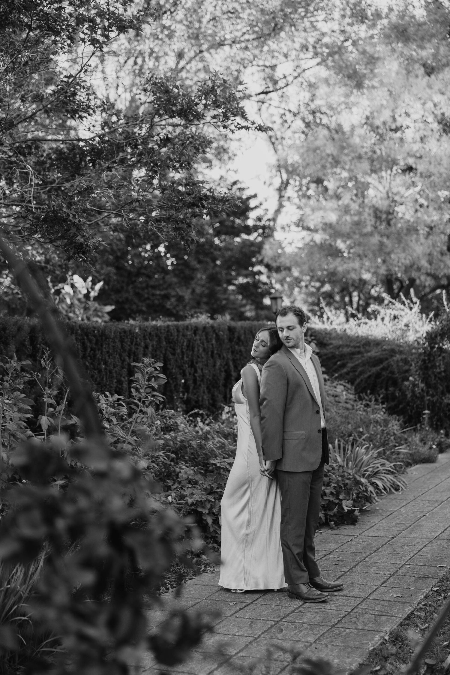 A black and white photo of a couple walking along a garden pathway lined with trees.