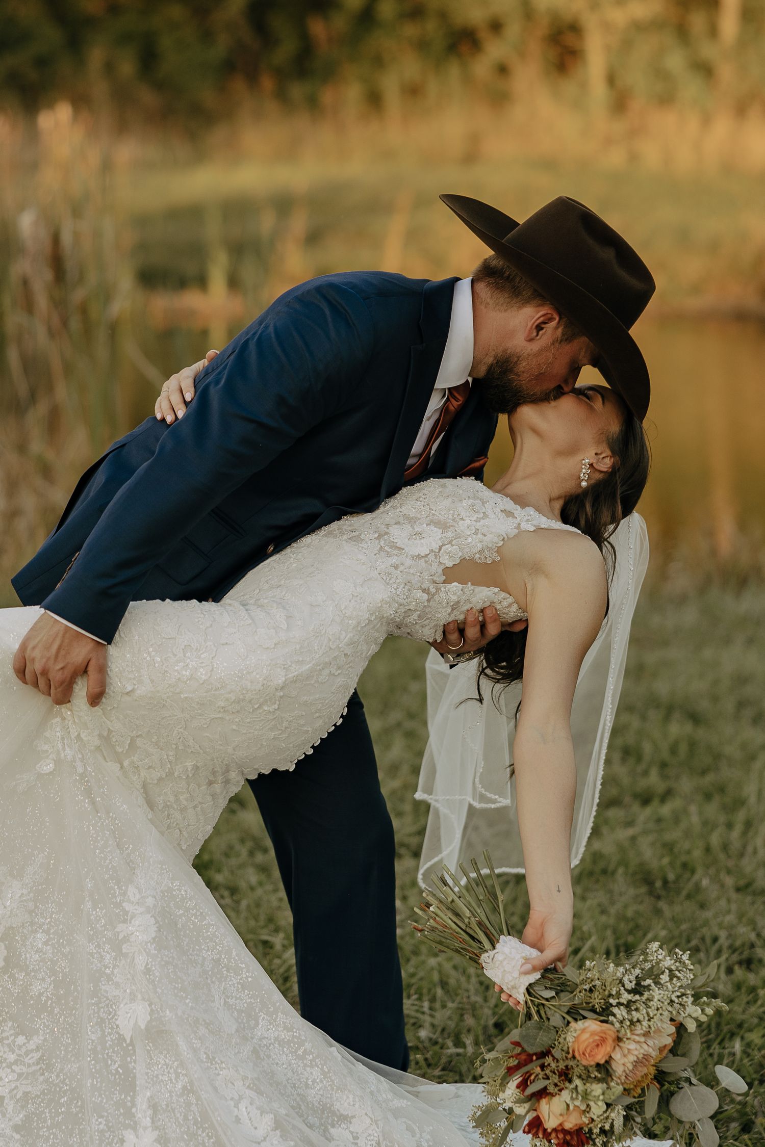 Romantic dip kiss moment between newlyweds by a lake during golden hour.