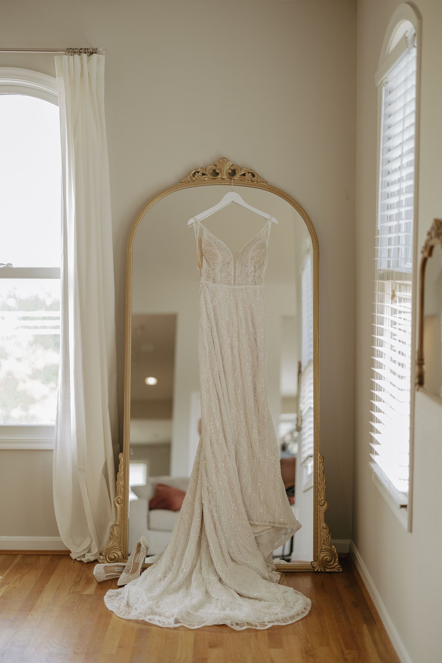 A white lace wedding dress hangs elegantly in a sun-lit room with white walls and hardwood floors.