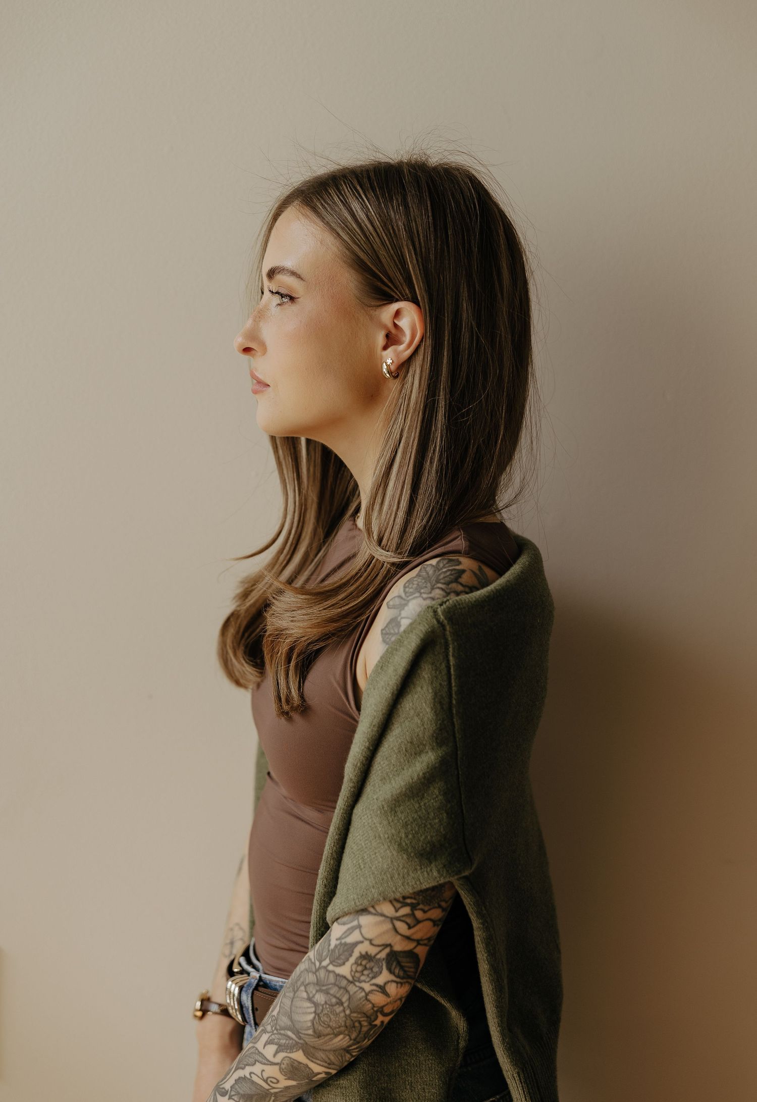 Side profile of a young woman with tattoos wearing a green jacket against a neutral background.