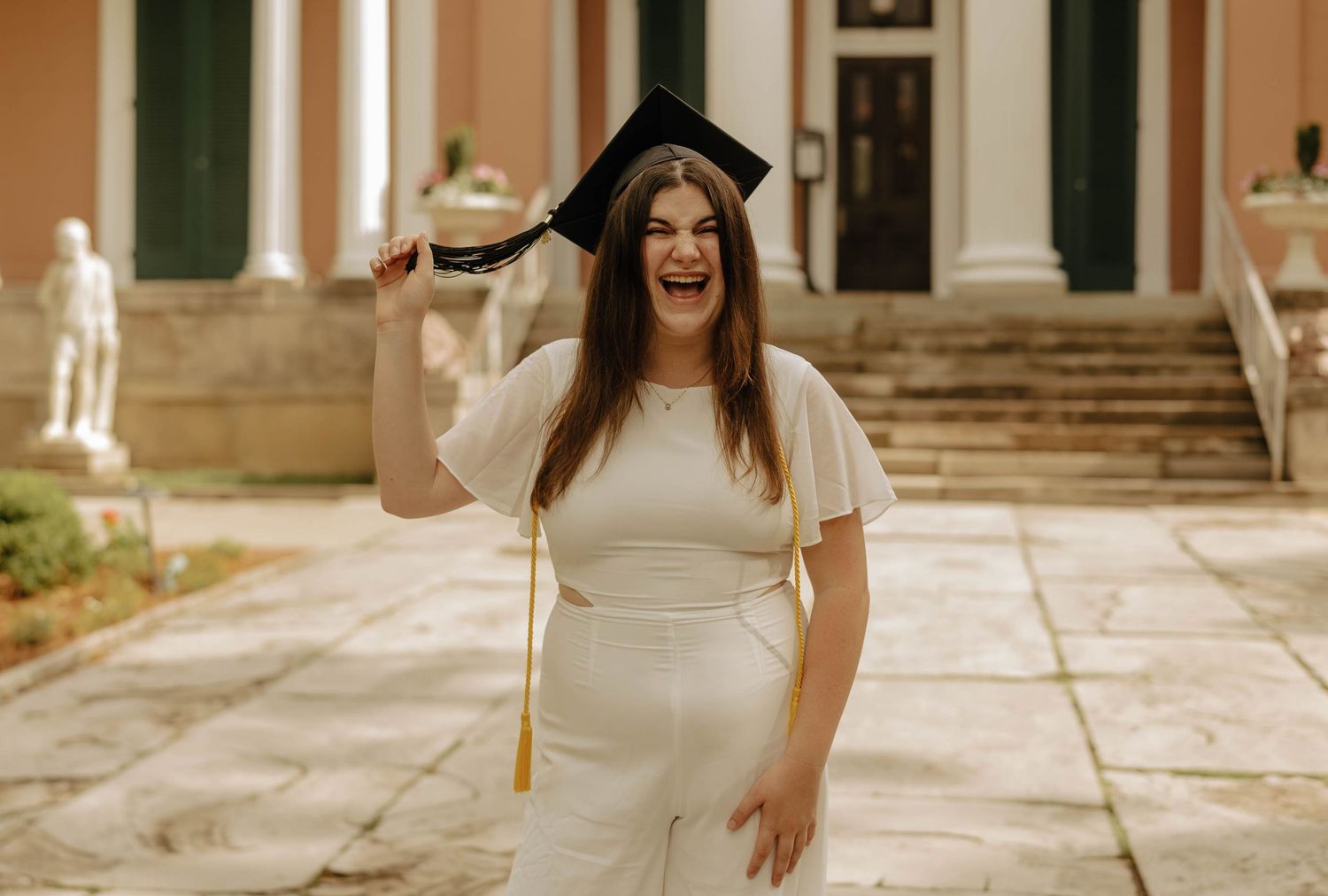 A graduate in a white dress celebrates joyfully on stone steps of a historic building.