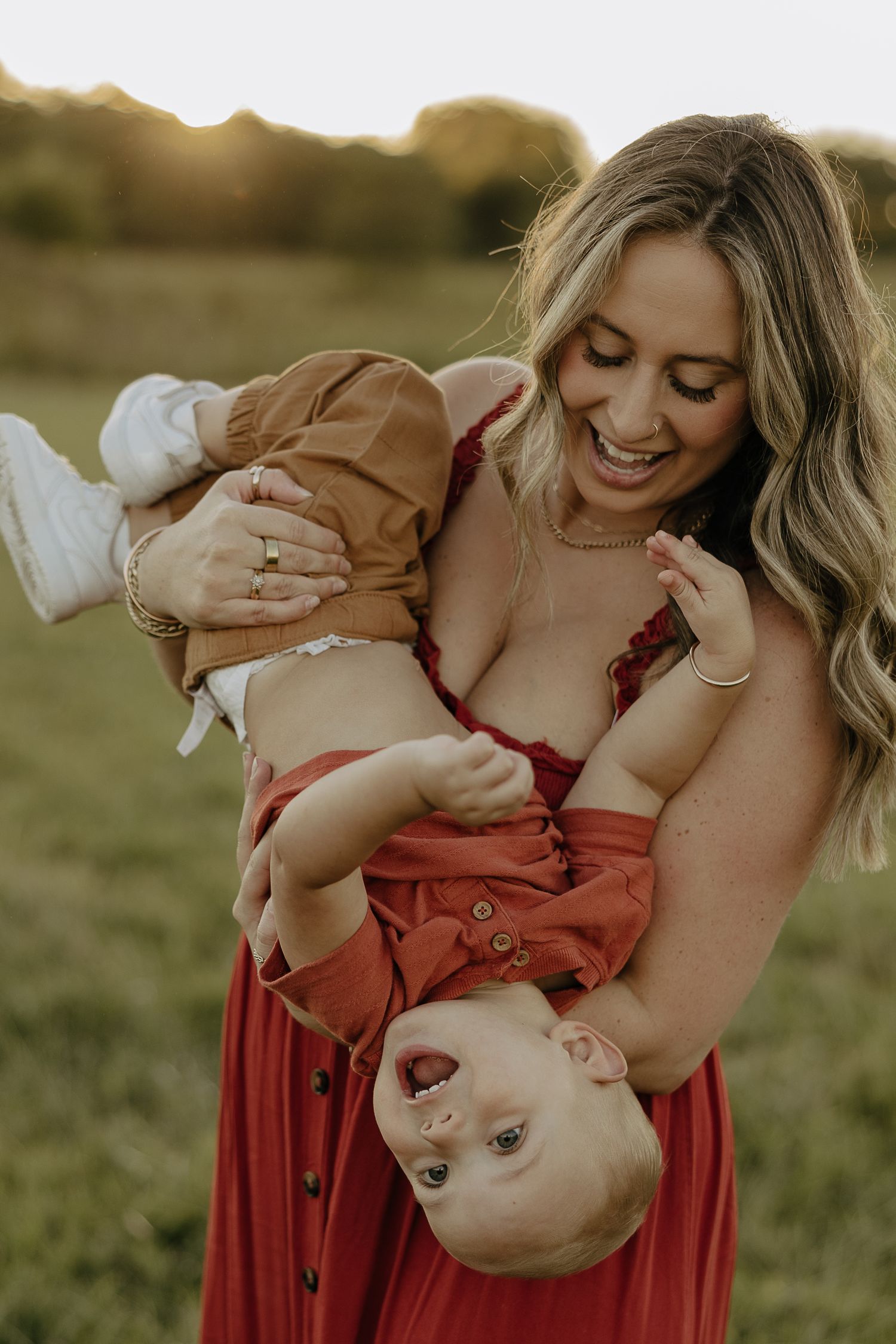 Mother in red dress plays with baby outdoors during golden hour sunset photoshoot.