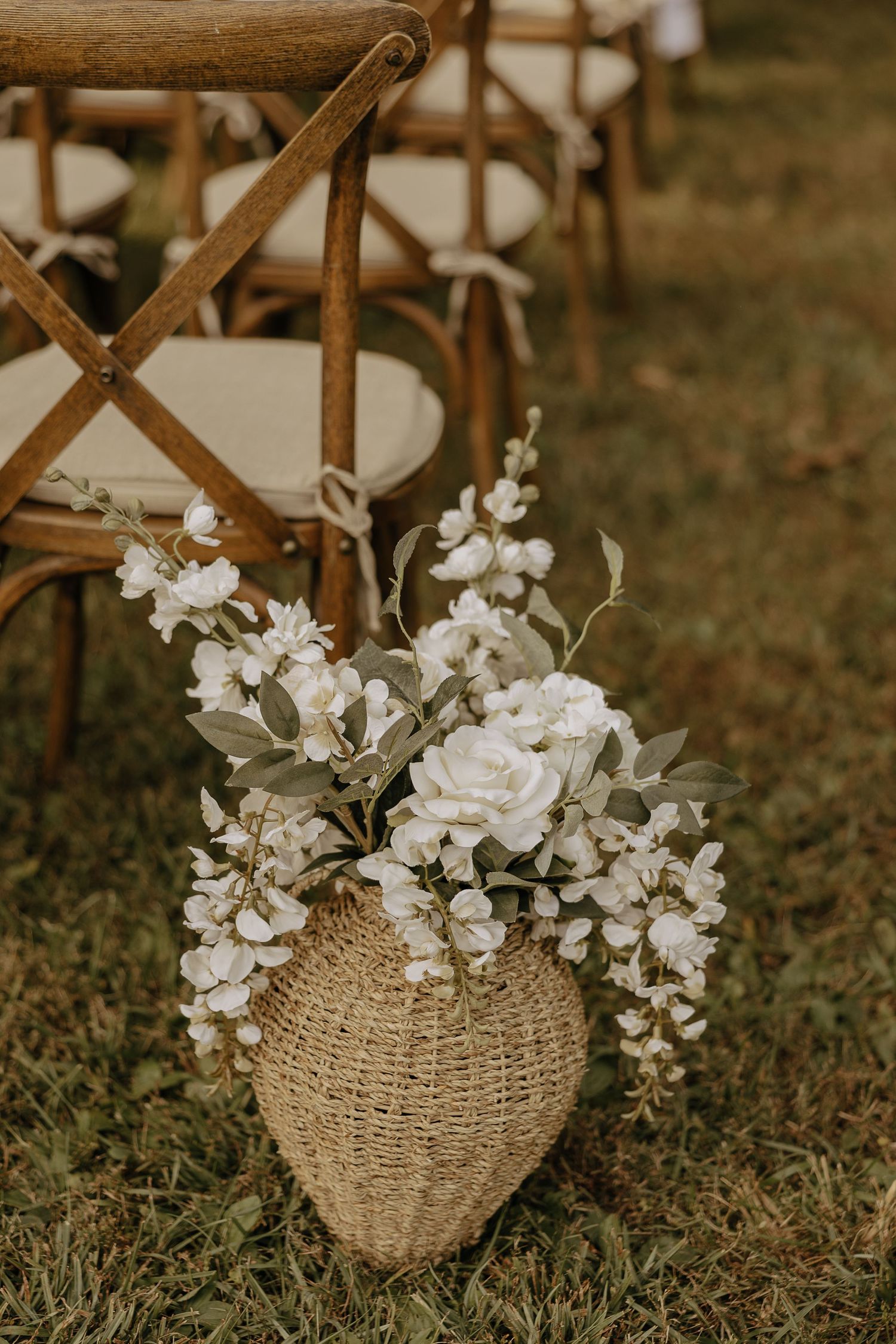 Woven basket with white floral arrangements placed on grass at outdoor wedding ceremony.