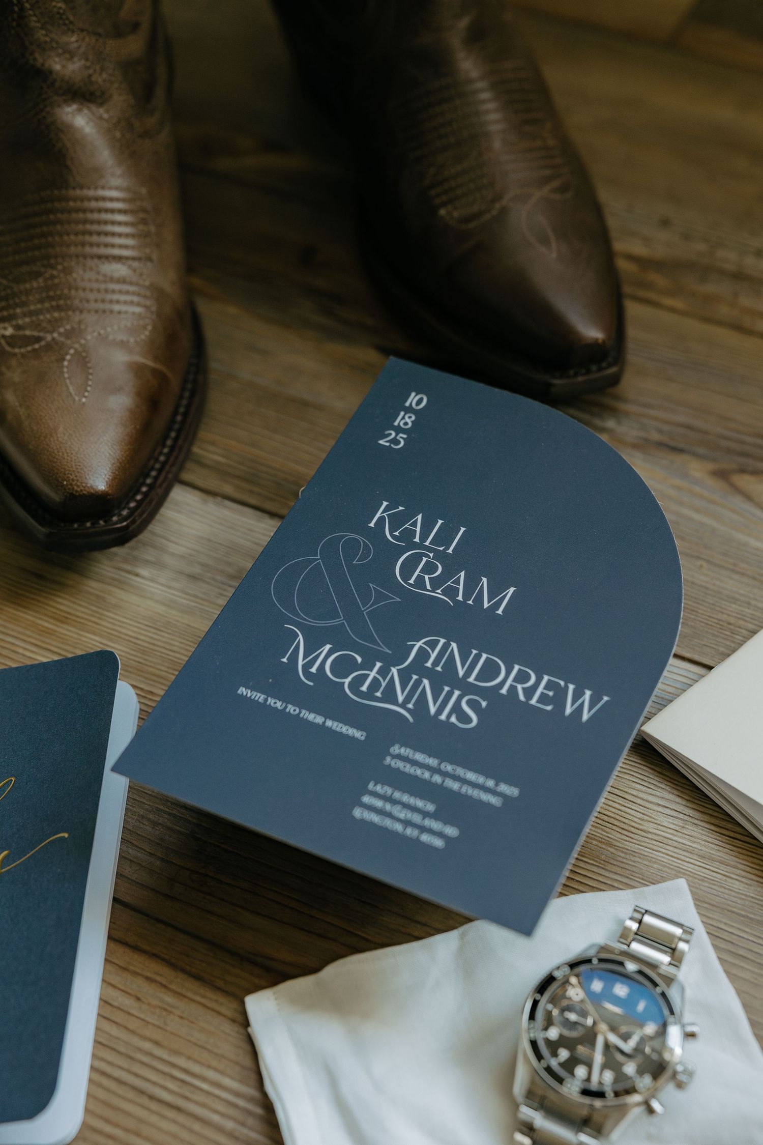 Wedding invitation and brown leather boots with a luxury watch on a wooden surface.