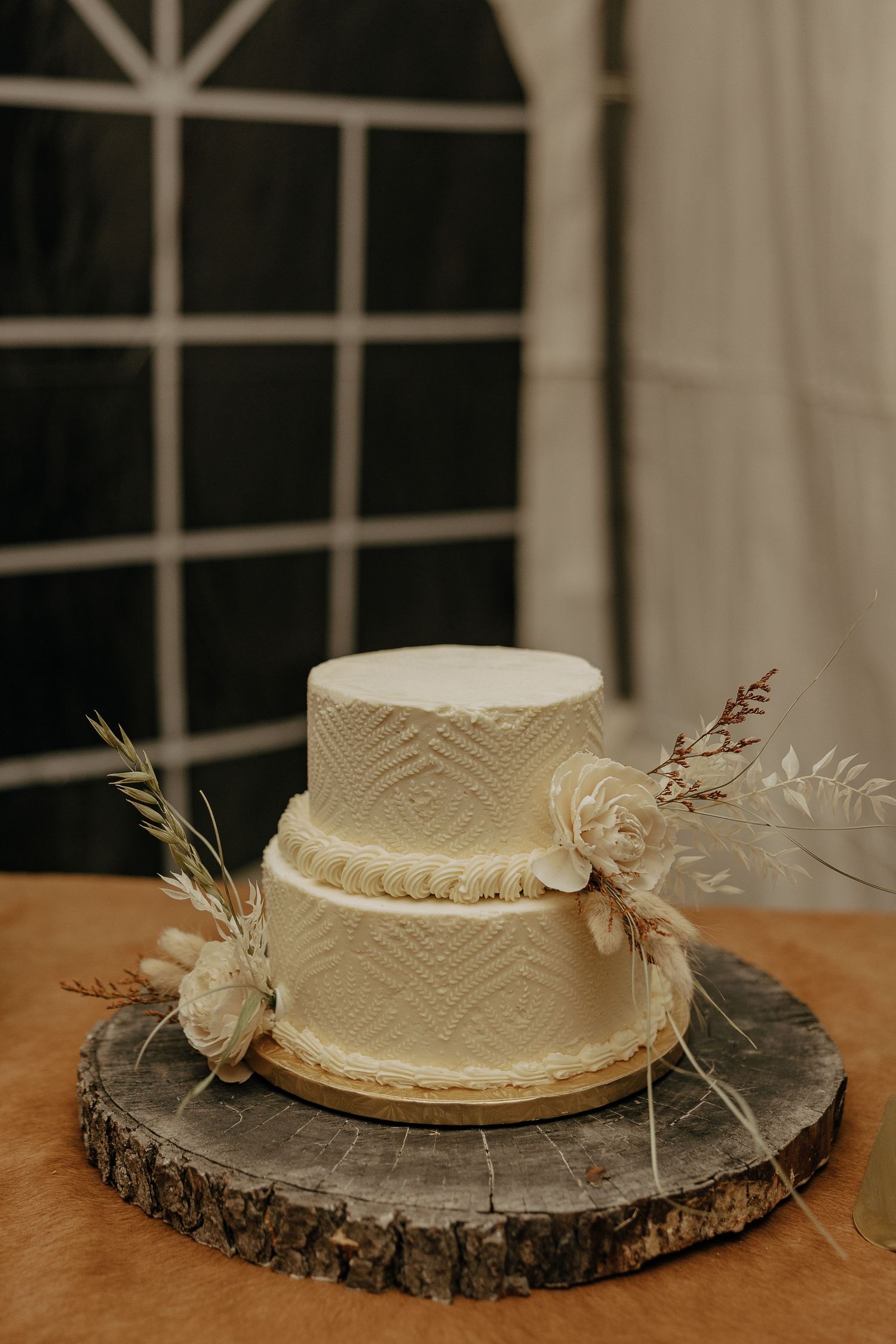 White wedding cake displayed on rustic wood slice with delicate lace patterns and dried floral accents.