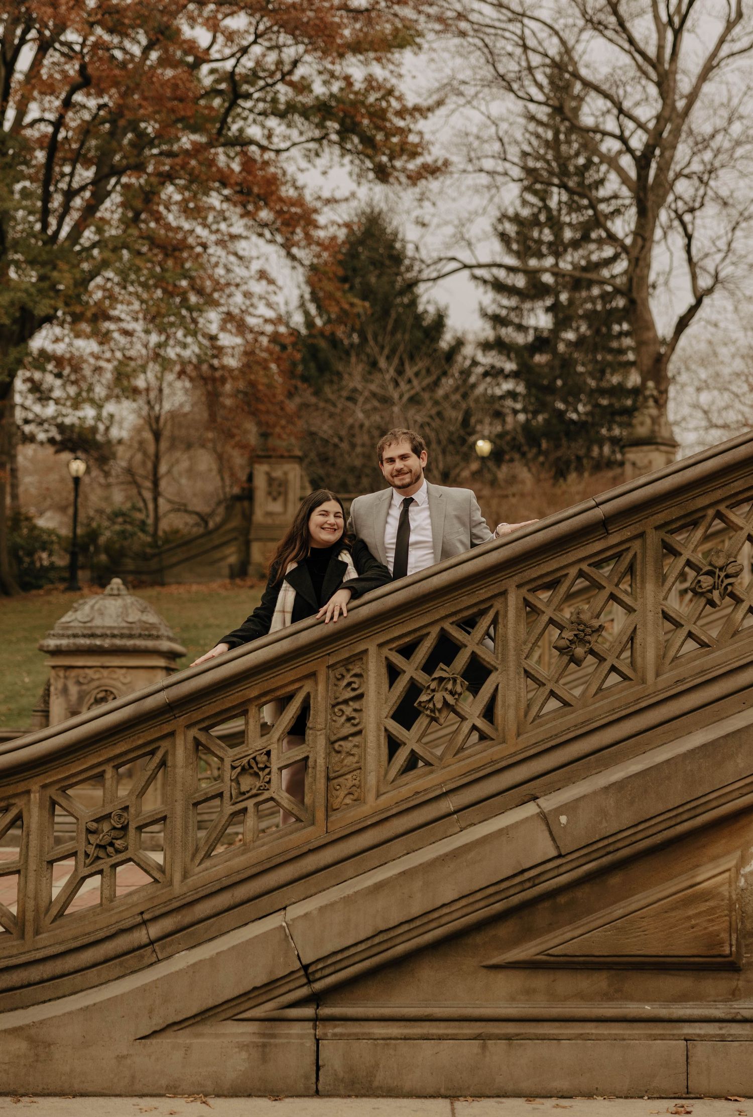 A couple stands together on ornate stone stairs in a park during autumn.