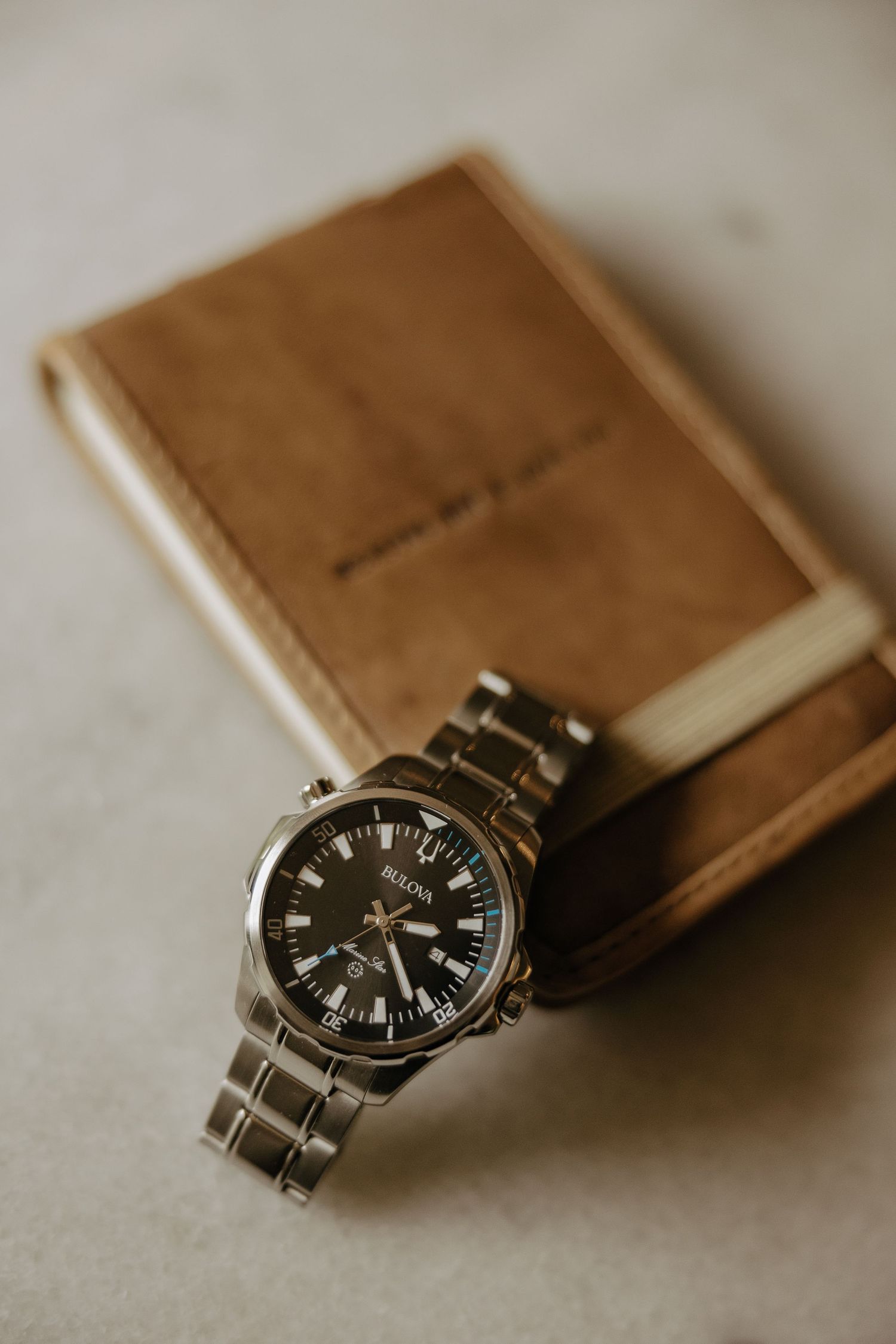 A silver watch rests on a brown leather wallet against a neutral background.
