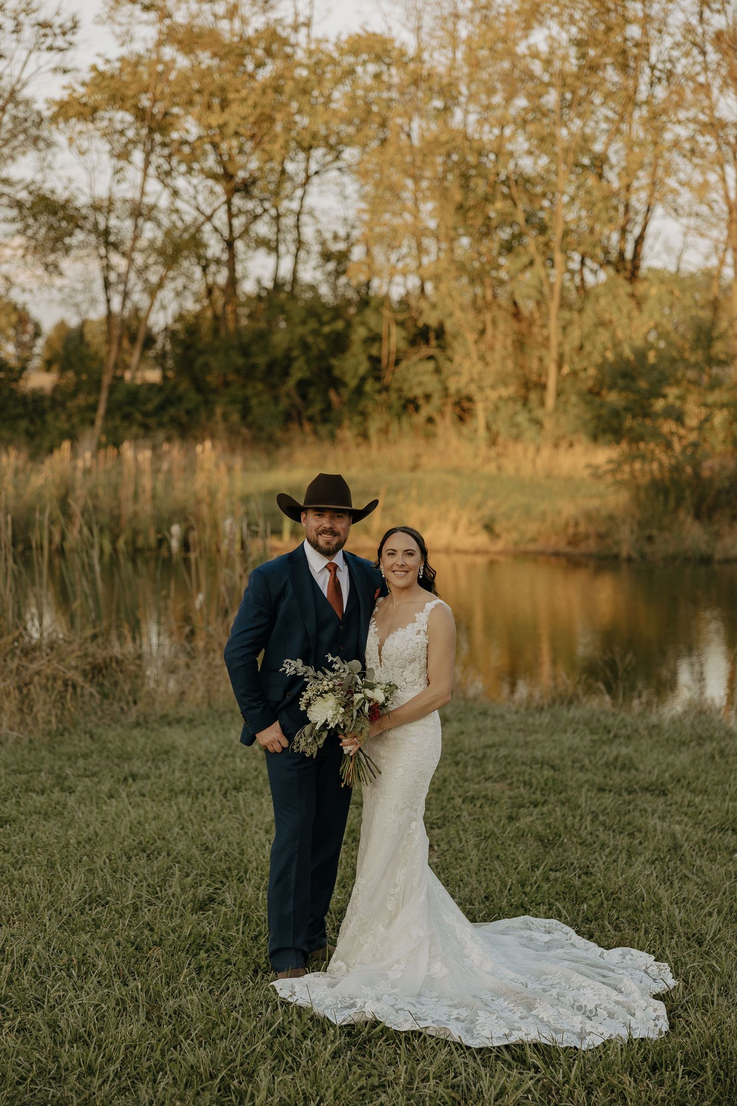 A romantic wedding portrait of a couple in western attire standing by a lake at sunset.