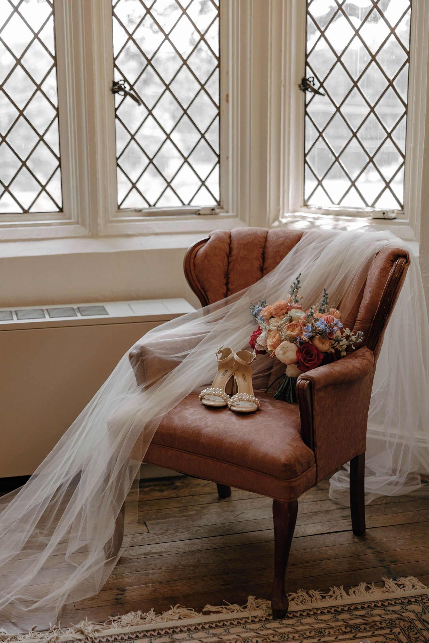 A flowing white wedding veil drapes elegantly over a vintage brown leather chaise lounge near diamond-paned windows.