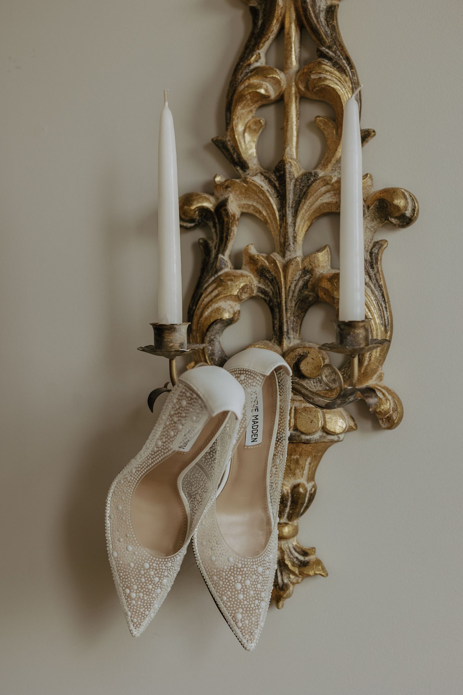 Wedding shoes displayed on a gold ornate wall sconce with candles creating a romantic bridal detail shot.
