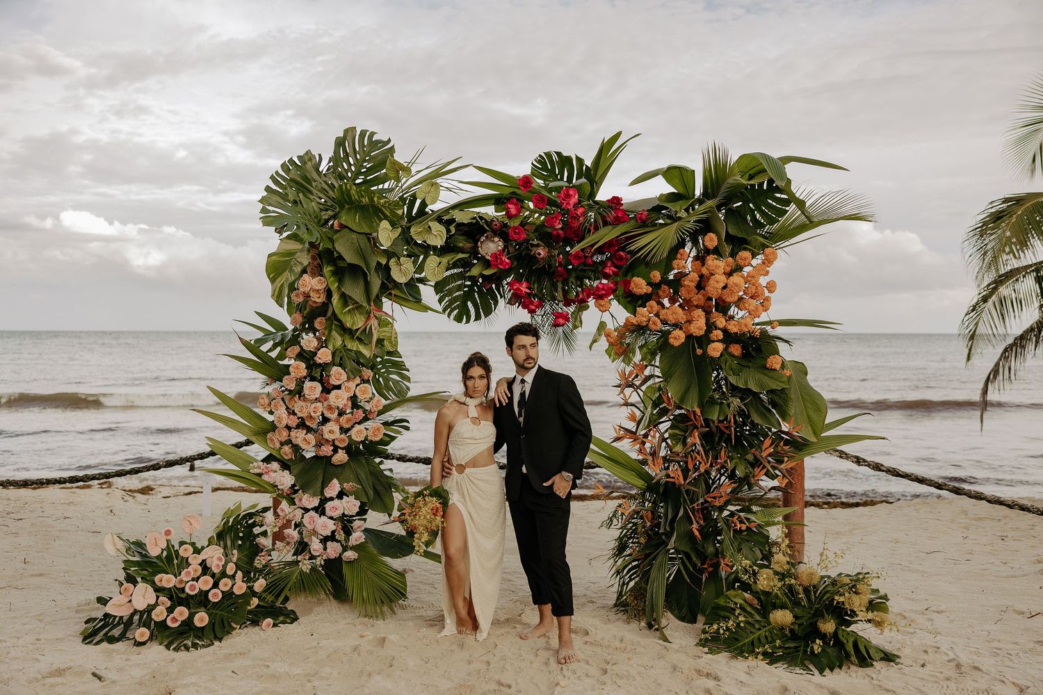A beachside wedding ceremony setup with tropical floral arrangements and an arch against a cloudy ocean backdrop.
