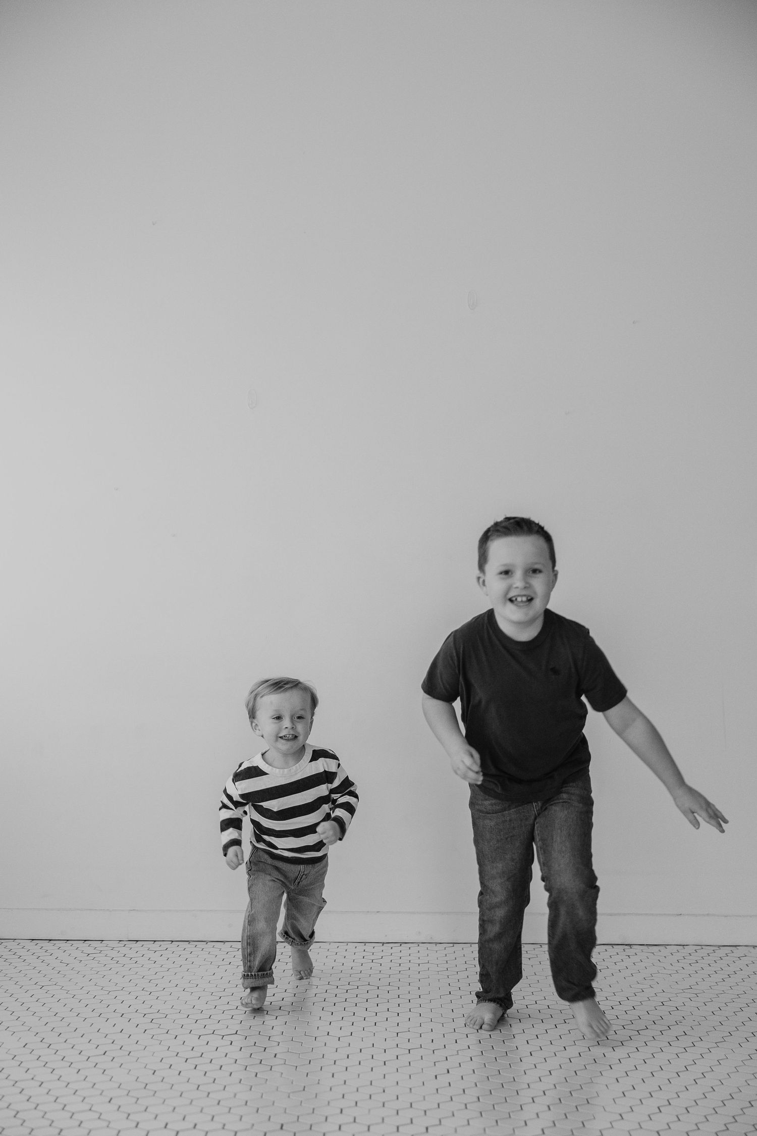 Two children run and play together against a minimalist gray wall background in black and white.