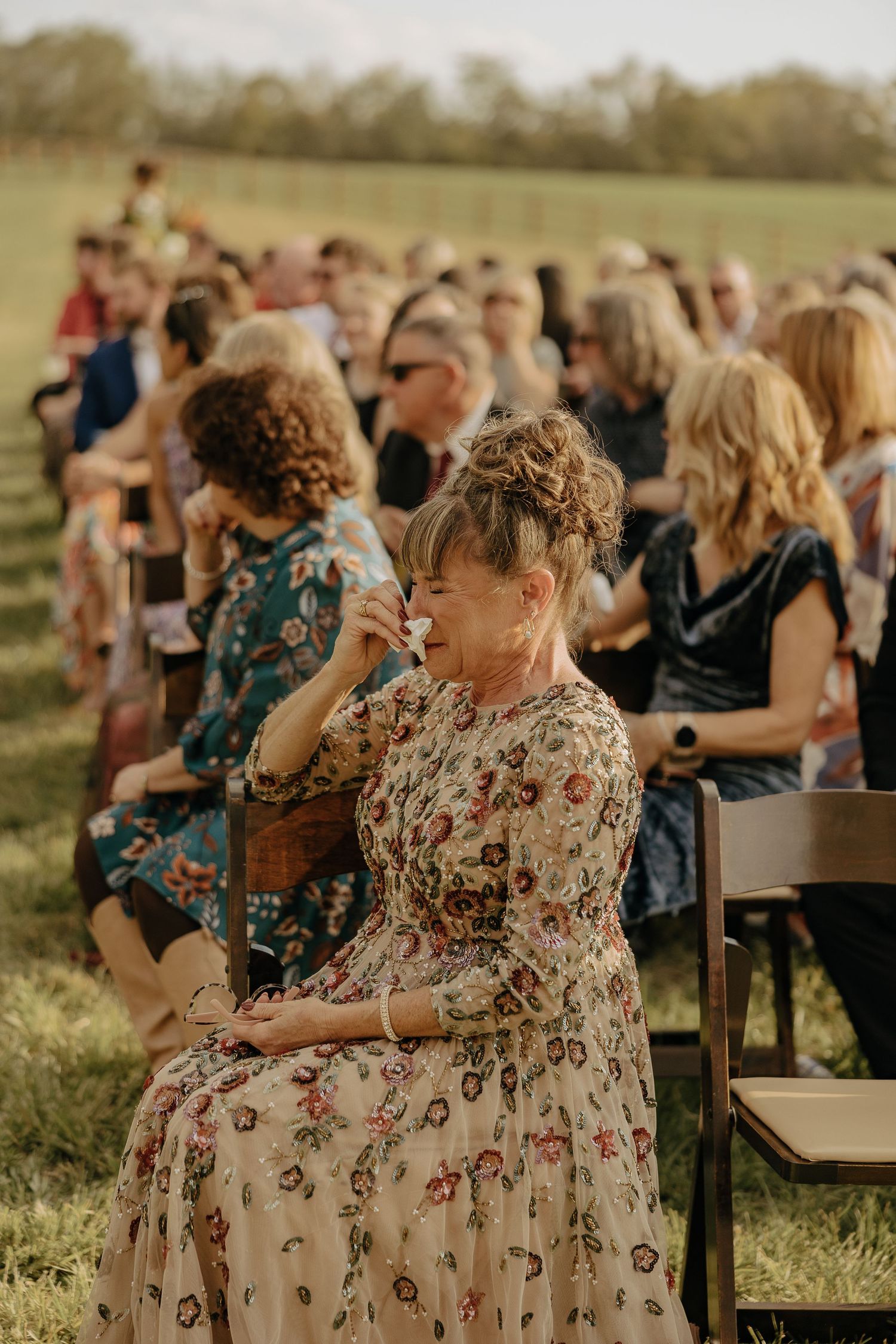 Wedding guests seated outdoors in floral dresses watching ceremony.