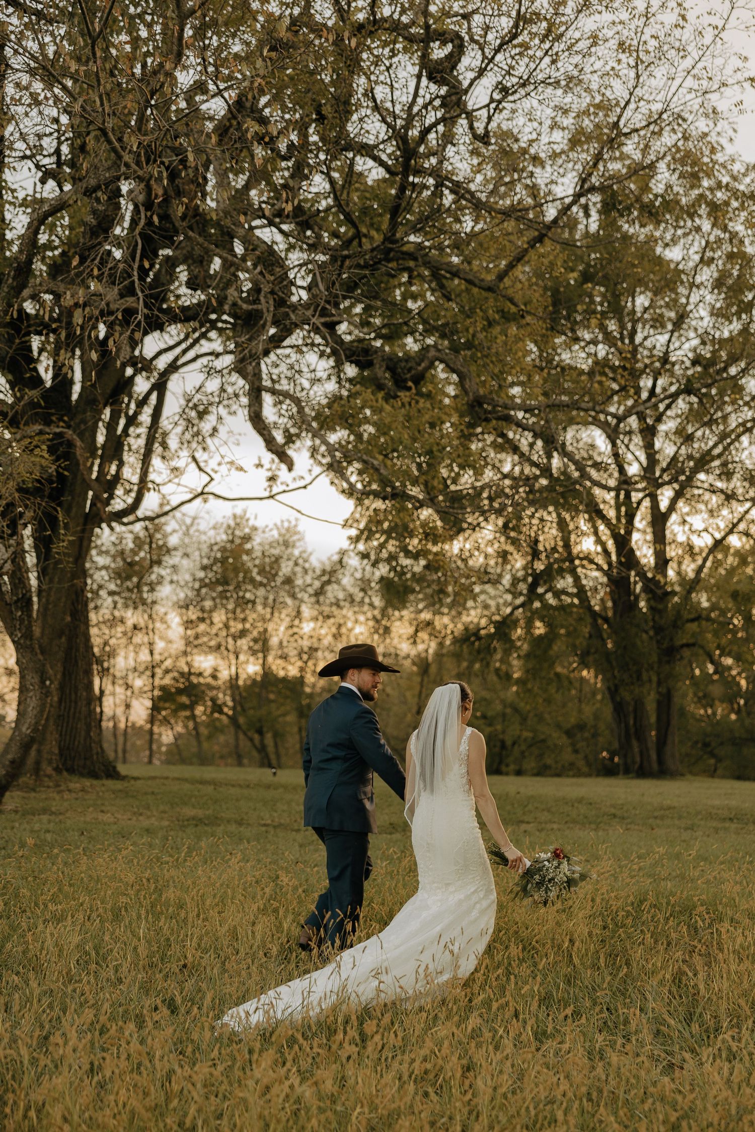 Wedding couple walks through golden field at sunset with trees in background.